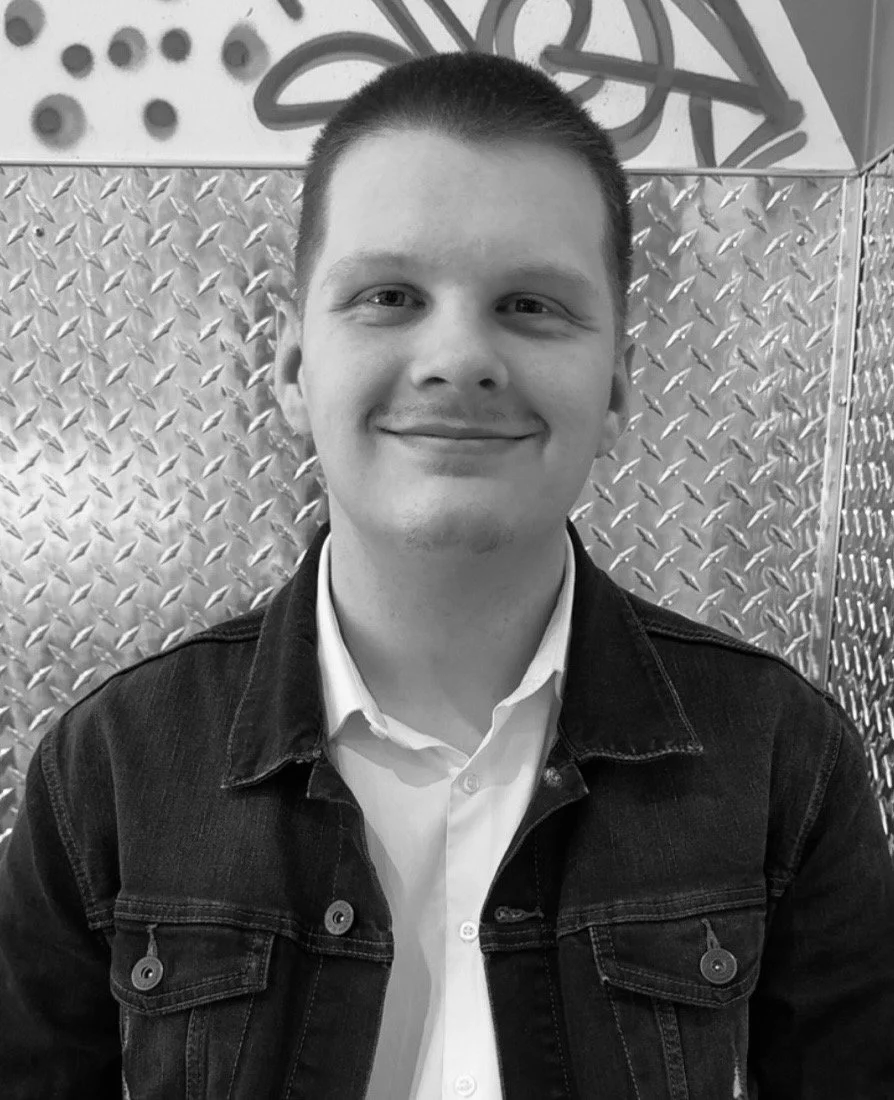A smiling young man with short hair, wearing a white shirt and dark jacket, standing in front of a metallic background with geometric patterns.