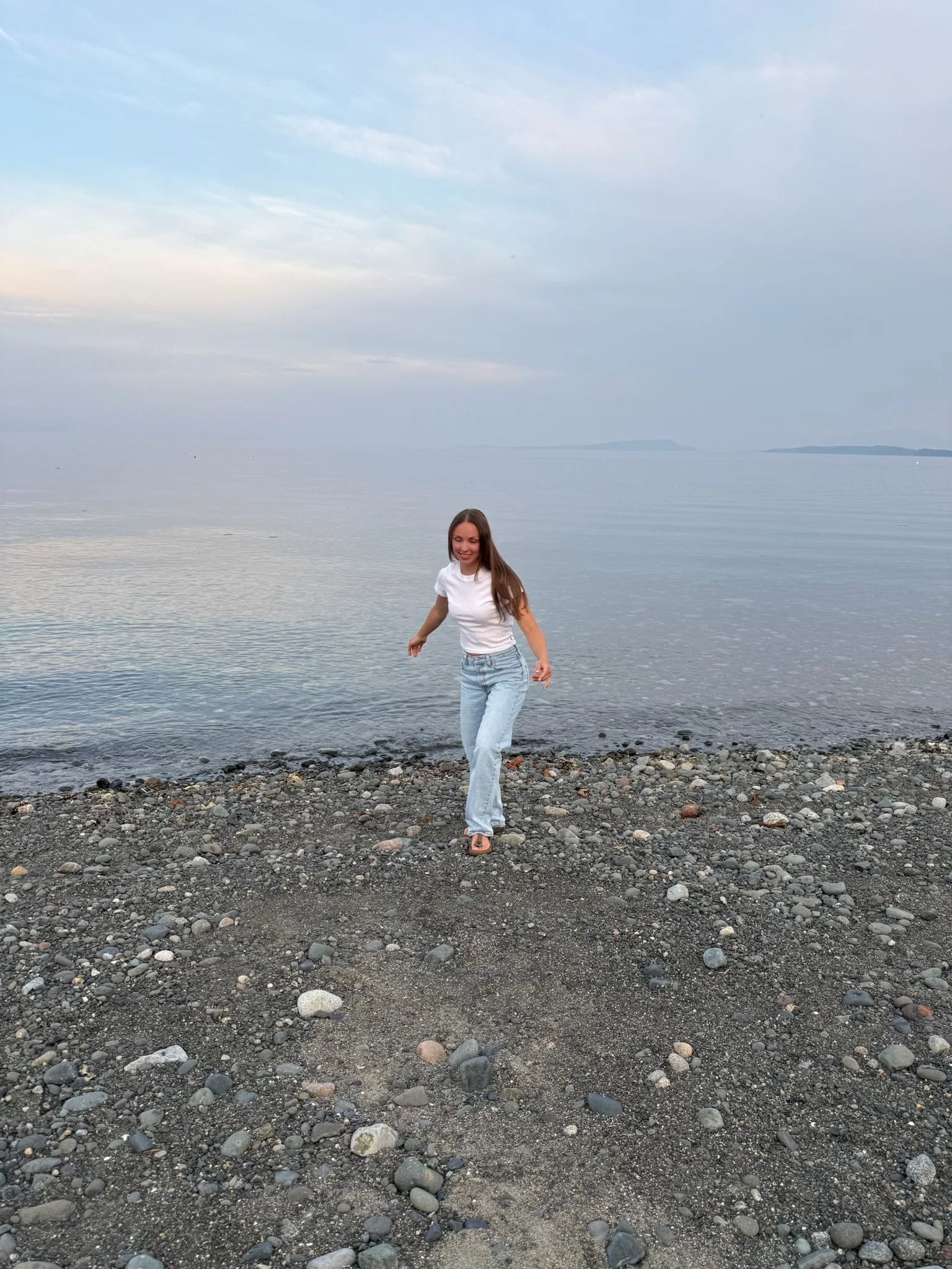 Woman walking on rocky beach near water with distant land and a cloudy sky.