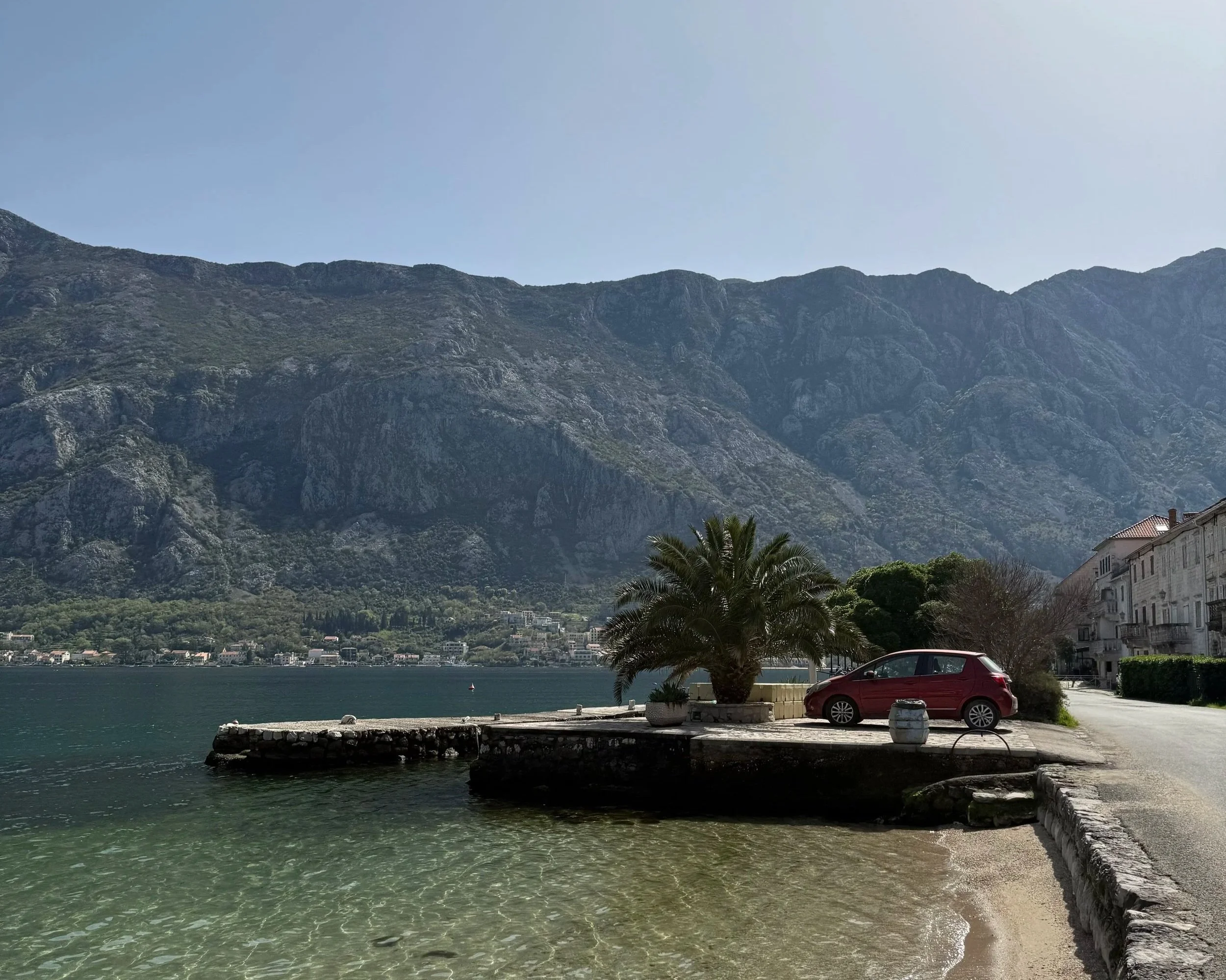 A scenic lakeside view with mountains in the background, a clear blue sky, a small pier with a car parked nearby, and a palm tree in the foreground.