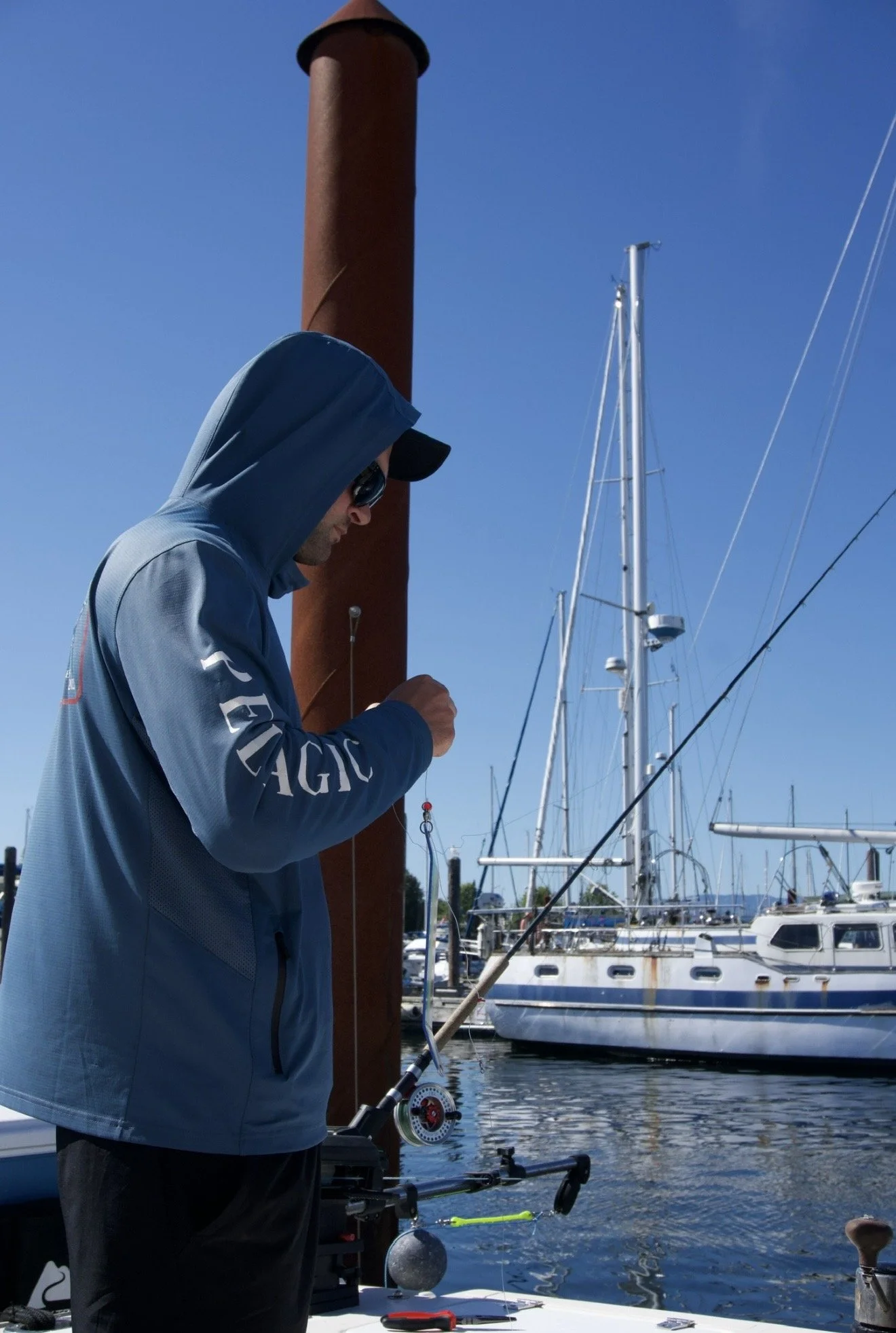 A man in a blue hoodie and sunglasses fishing at a marina on a clear day with several sailboats docked in the background.