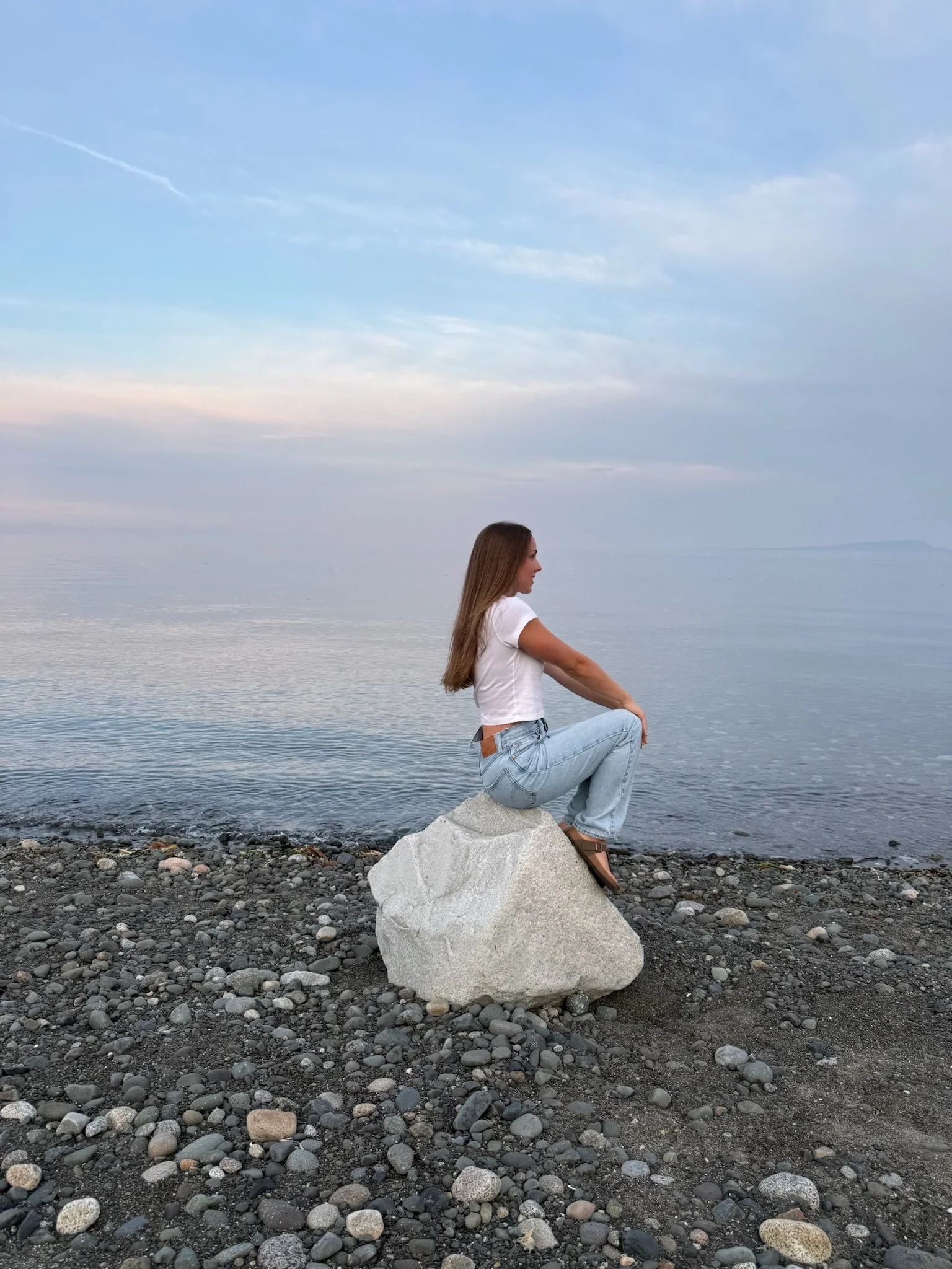 A woman with long hair sitting cross-legged on a large rock by a pebbled shoreline, facing to the right, with a calm body of water and a cloudy sky in the background.