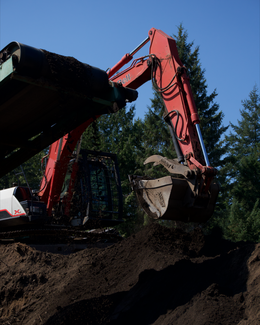 A red excavator digging into the dirt with a forest of evergreen trees in the background under a clear blue sky.