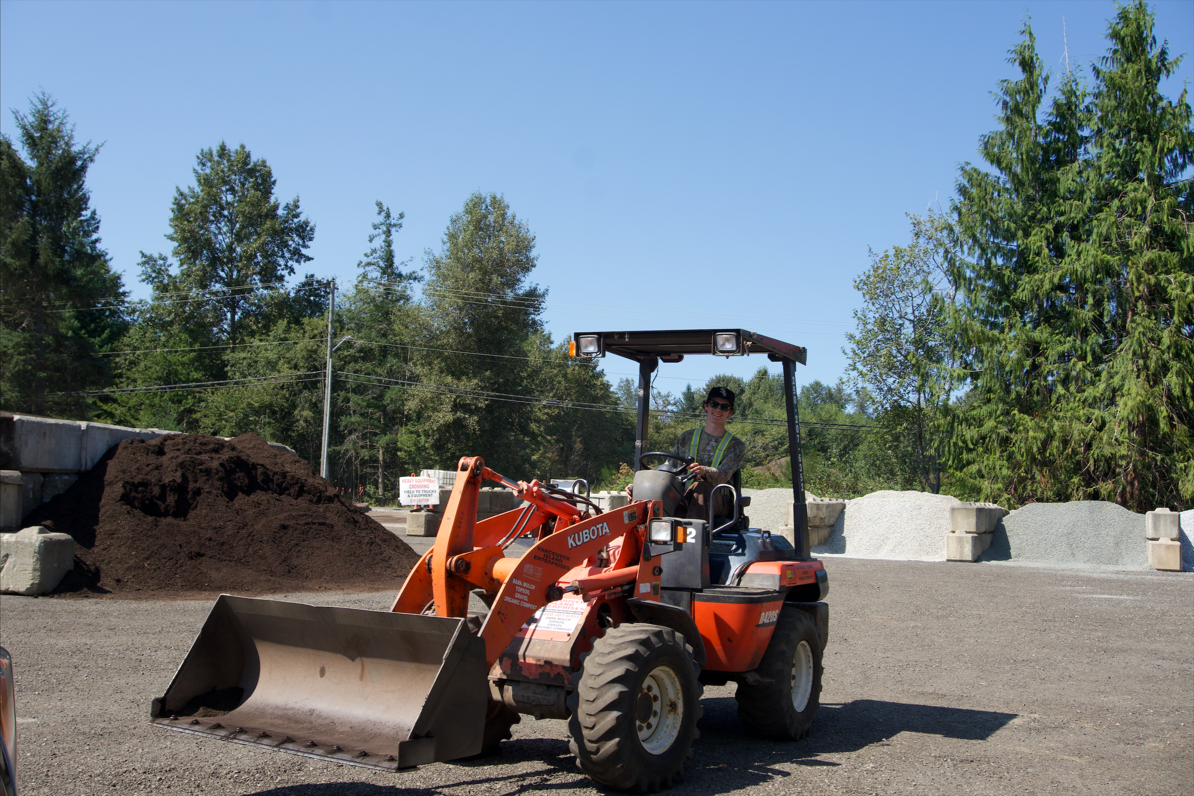 A person operating an orange Kubota compact construction loader on a gravel surface surrounded by trees and construction materials.