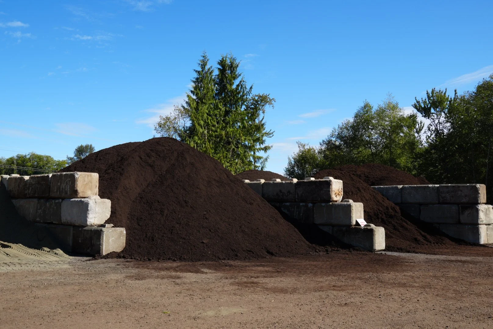 Mounds of dark soil behind stacked concrete blocks in an outdoor area with trees and a blue sky.