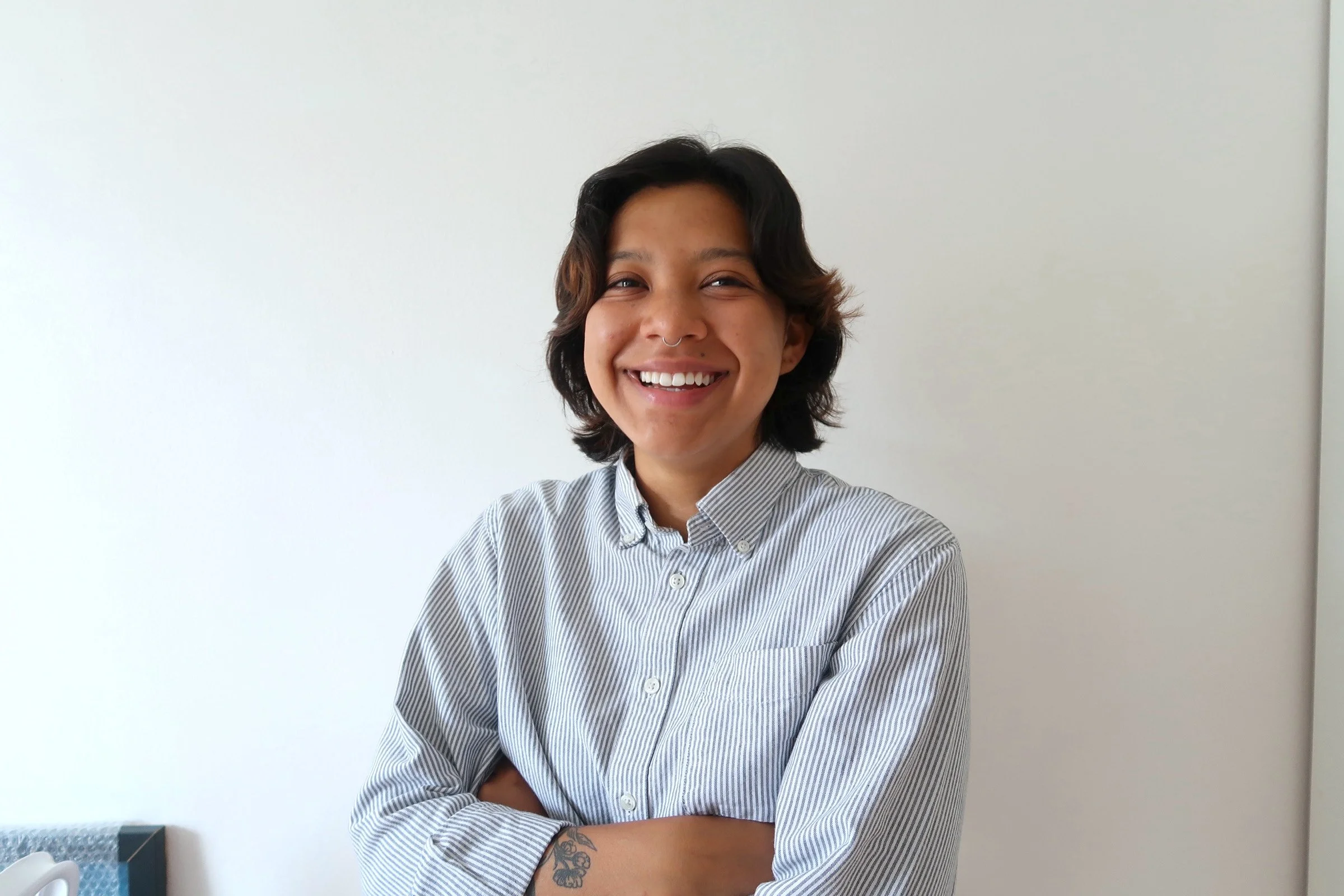 A young person with short, wavy dark hair, smiling and looking at the camera, wearing a striped button-up shirt, standing in front of a white wall.