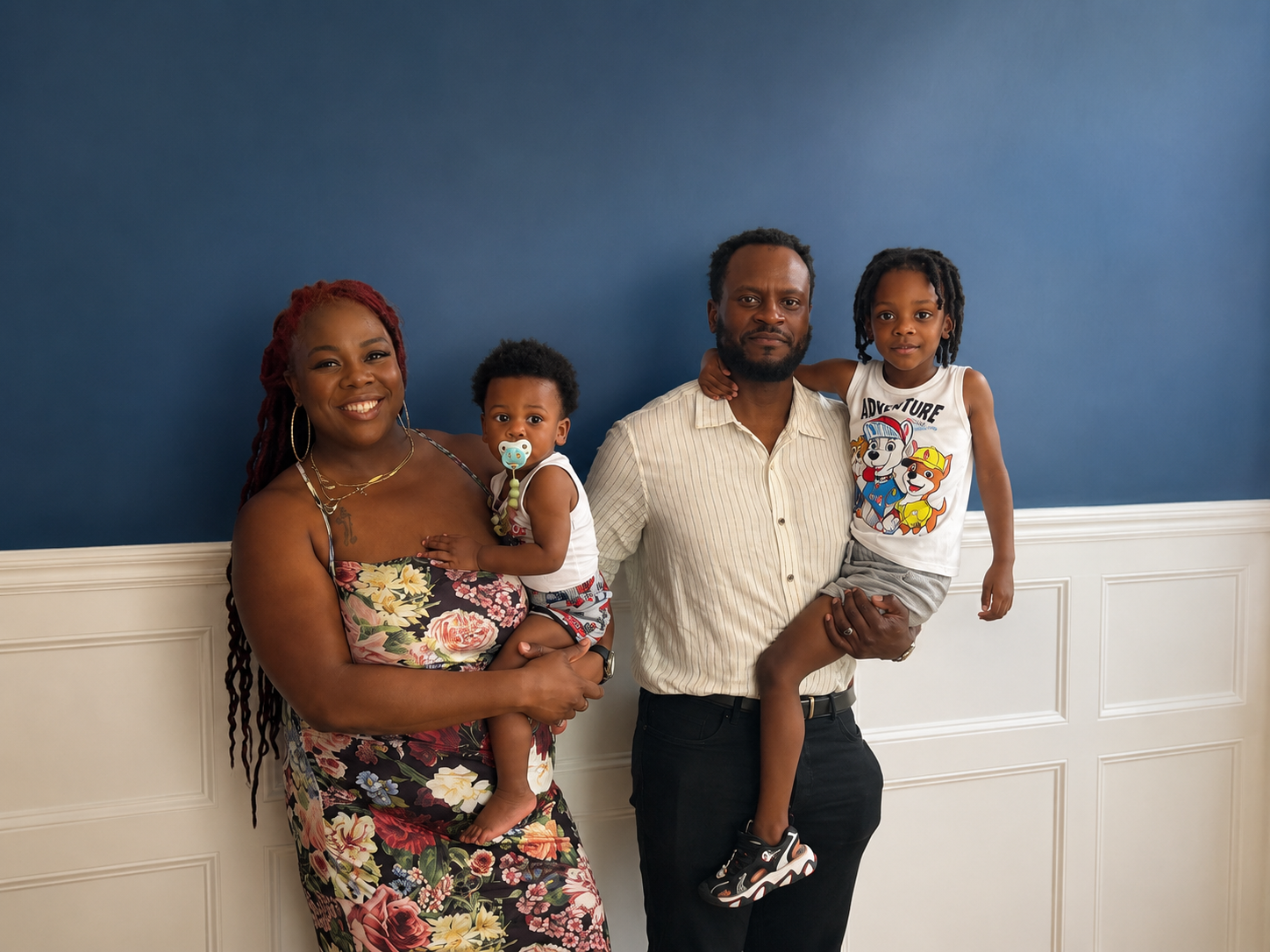 A family of four standing against a blue wall, with the mother on the left wearing a floral dress and the father on the right wearing a light-colored striped shirt. The mother is holding a young child with a pacifier, and the father is holding an older child wearing a cartoon-themed tank top.