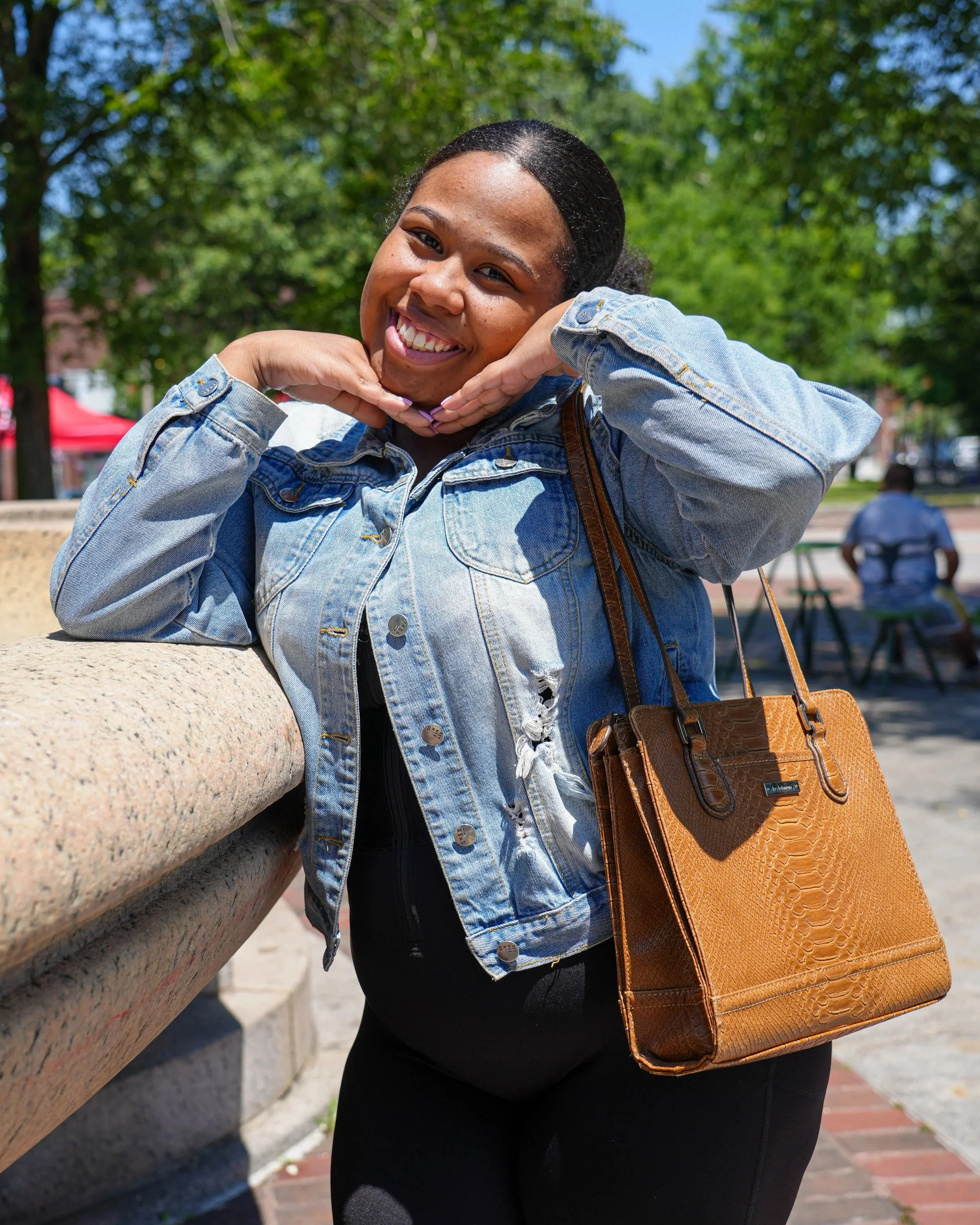 A woman with dark hair smiling and posing with her hands under her chin, wearing a denim jacket and a large brown handbag, outdoors on a sunny day with trees and a bench in the background.