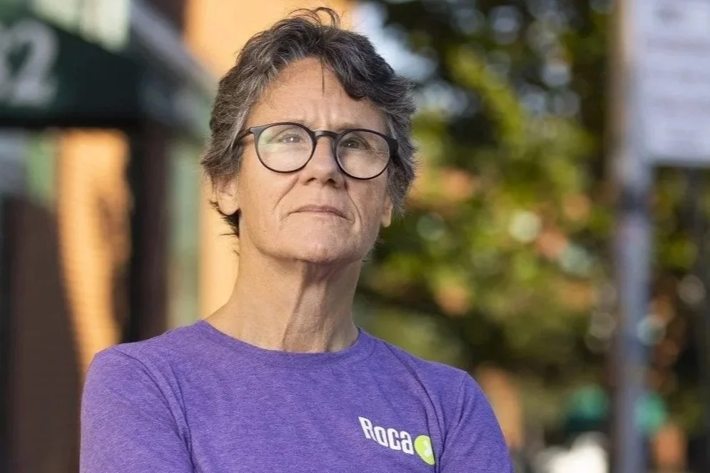 Woman with short, grey hair and glasses standing outdoors with a neutral expression, wearing a purple shirt.