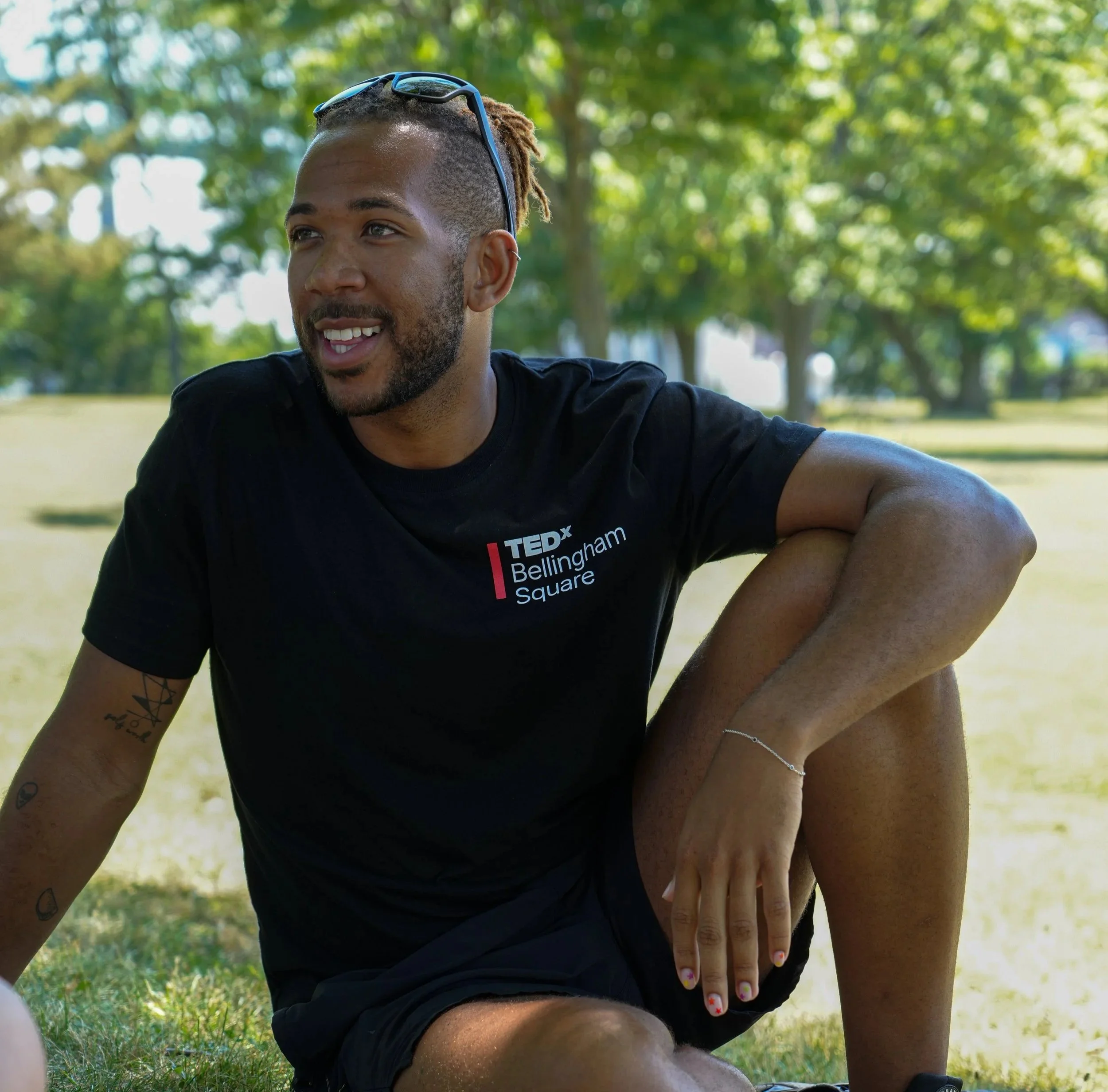 A smiling young man wearing sunglasses on his head, sitting outdoors in a park with green trees in the background. He is wearing a black T-shirt with white and red text.