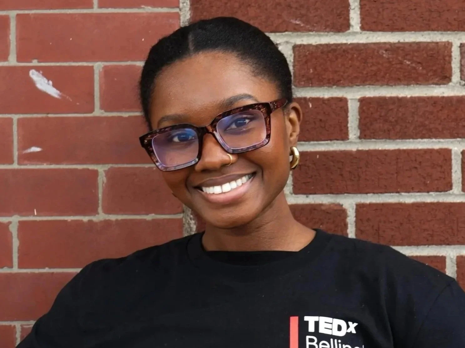 Young woman smiling, wearing large tortoiseshell glasses, earrings, a small nose ring, and a black TEDxBellingham Square shirt, standing against a red brick wall.