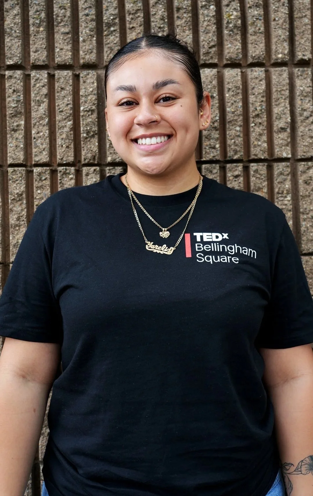 A woman with dark hair pulled back, smiling, wearing a black TEDx Bellingham Square t-shirt, layered necklaces, and earrings, standing in front of a brick wall.