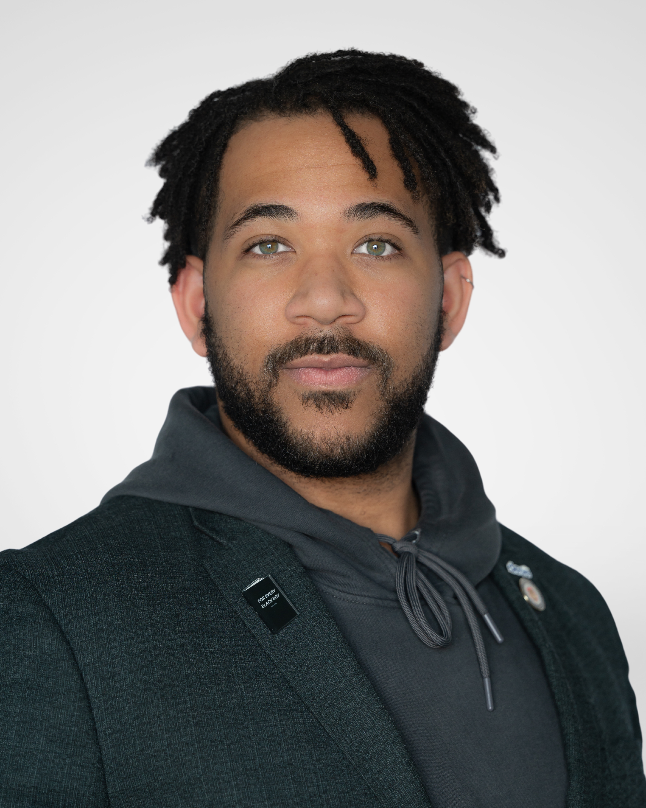 Close-up of a man with gorgeous deep brown skin, dark curly hair, hazel eyes, and a beard, wearing a black jacket and a dark gray hoodie, with a name tag that reads 'For Every Black Boy'.