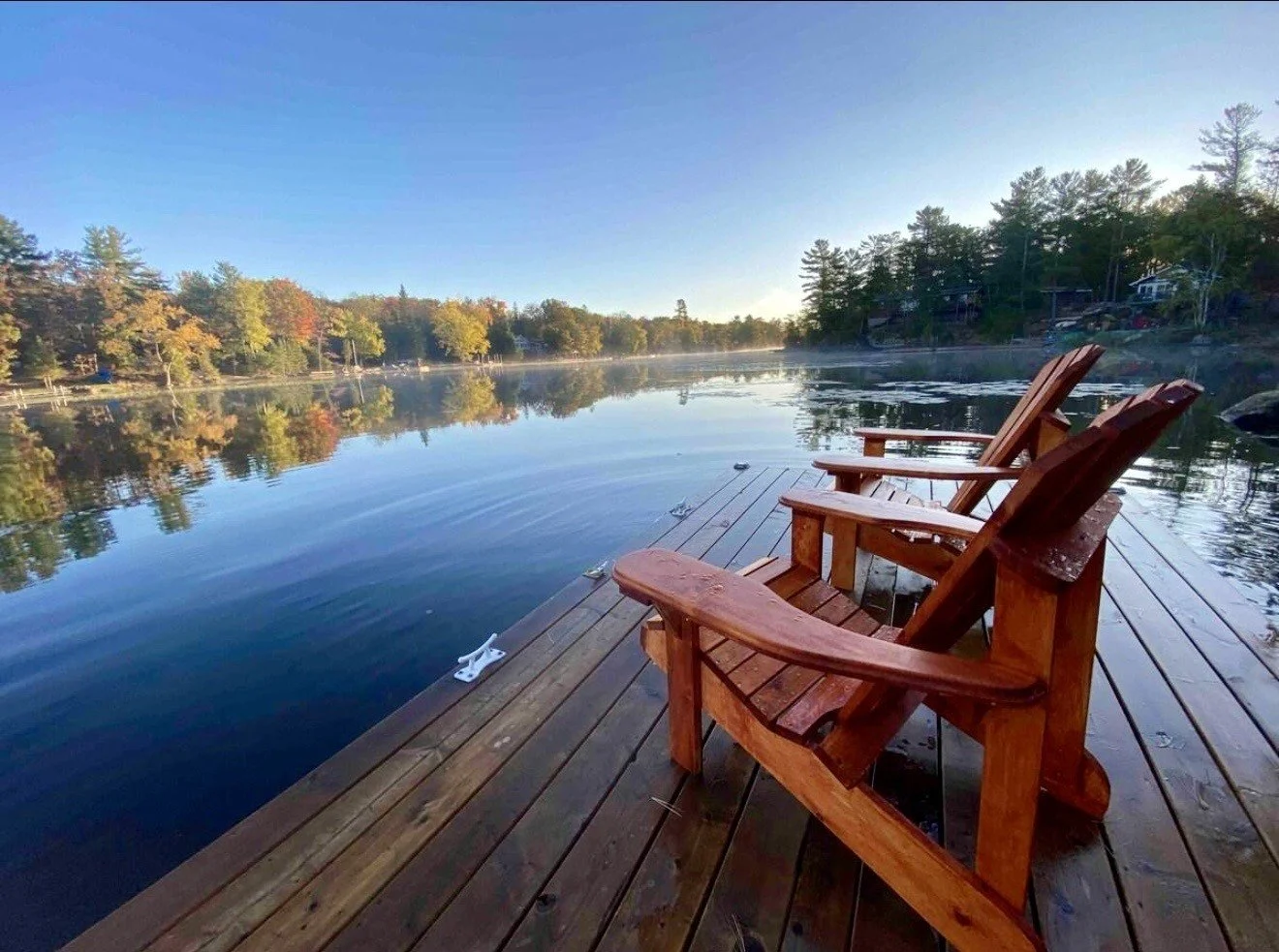Three wooden Adirondack chairs on a dock overlooking a calm lake with reflections of trees on the water, under a clear blue sky.