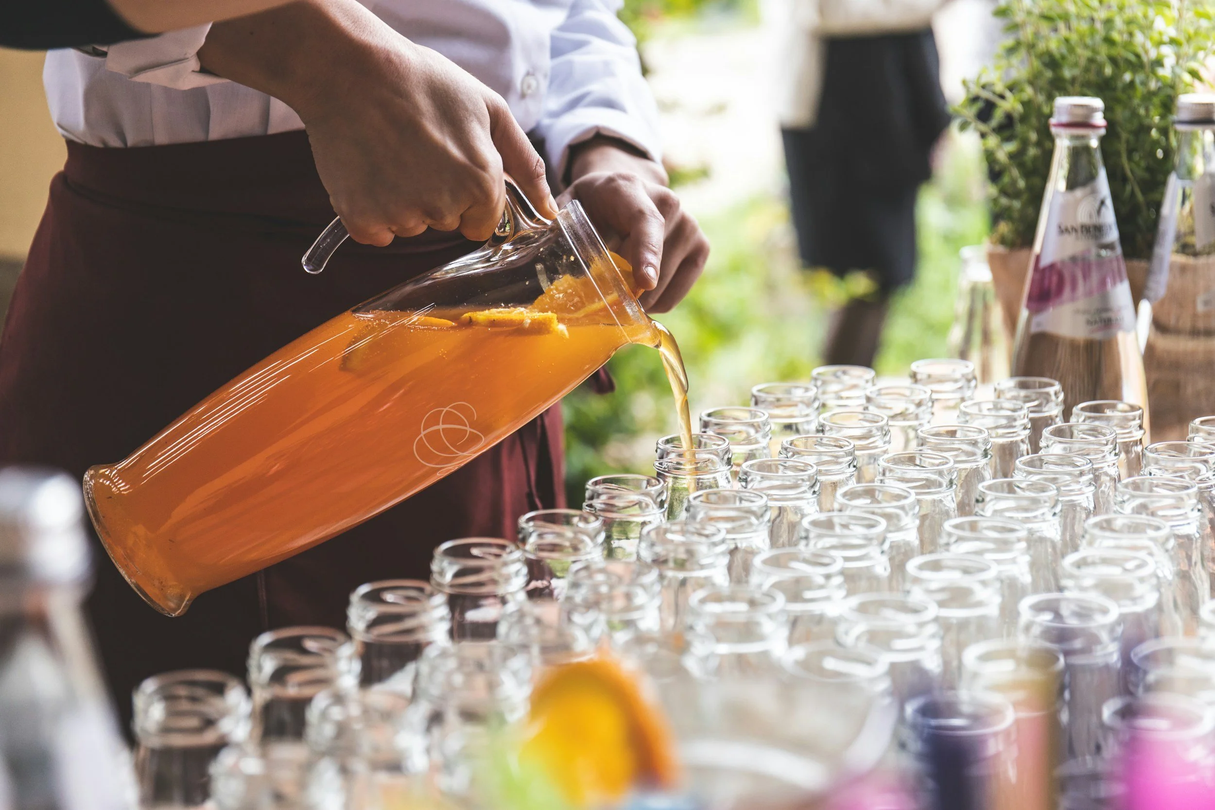 Person pouring orange juice into small glasses at an outdoor event with bottles and greenery in the background.