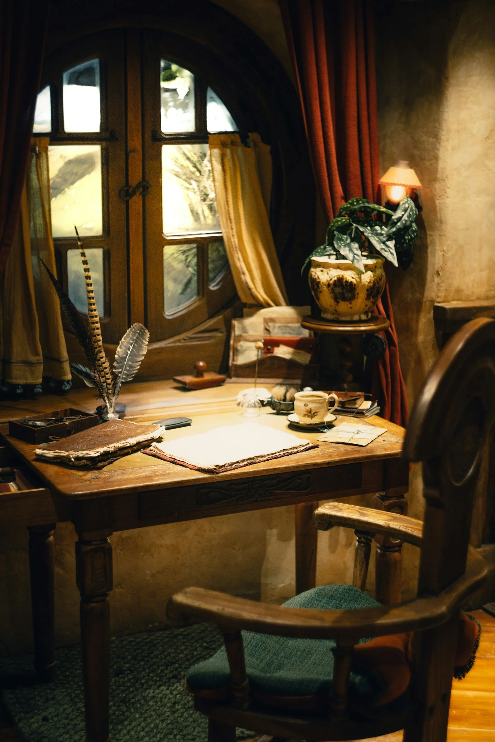 A cozy interior scene with a wooden table and chairs, a window with yellow curtains, a potted plant with large green leaves, a small lamp, and various objects including books, feathers, and a tea cup.