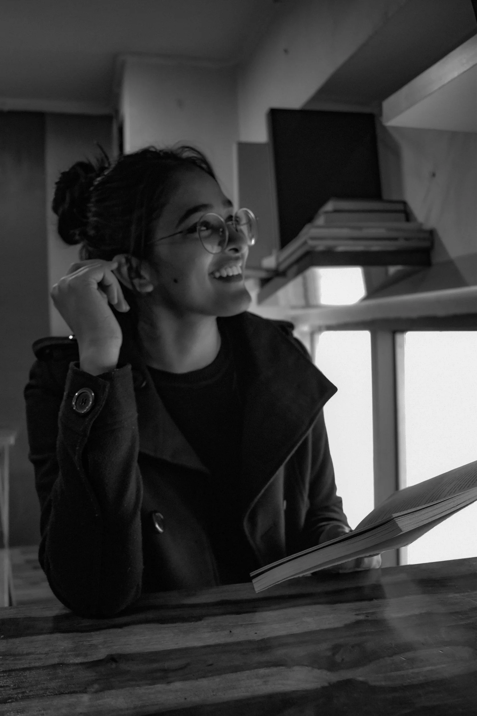 A young woman with glasses smiling while sitting at a table reading a book, in a room with a bookshelf and windows.