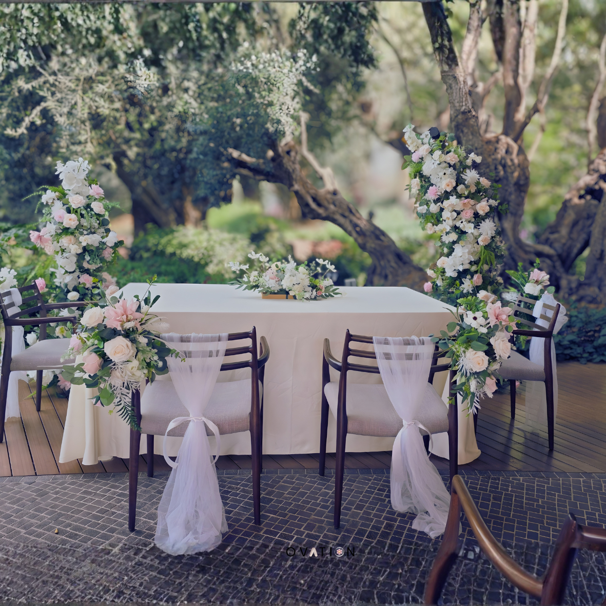 Wedding reception setup with a long table, decorated with floral arrangements and chairs draped with white fabric, set outdoors under trees.