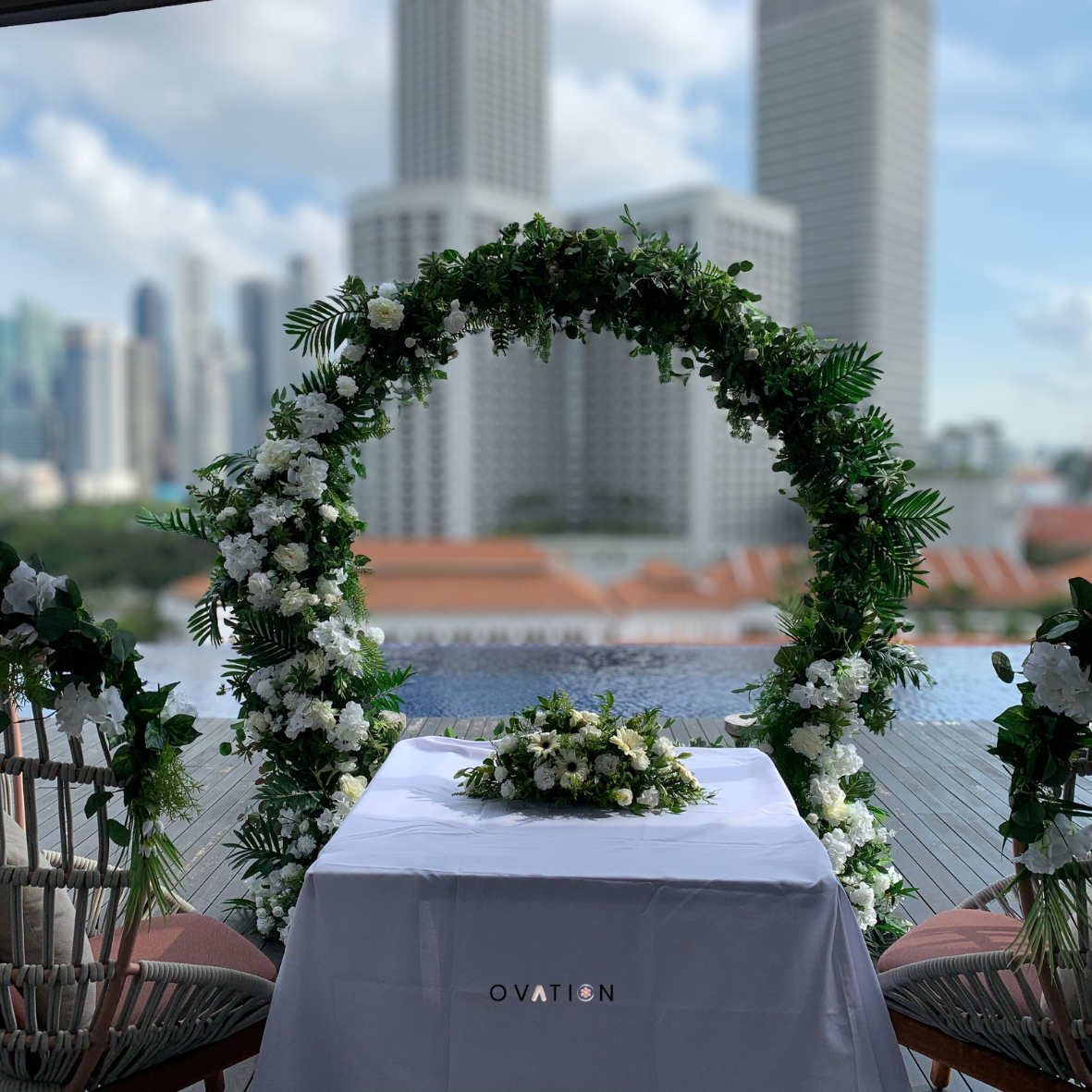 A wedding altar decorated with a large circular floral arrangement of white flowers and green foliage, with a bouquet of white flowers on a table covered with a white cloth, set against a cityscape background with tall buildings and cloudy sky.
