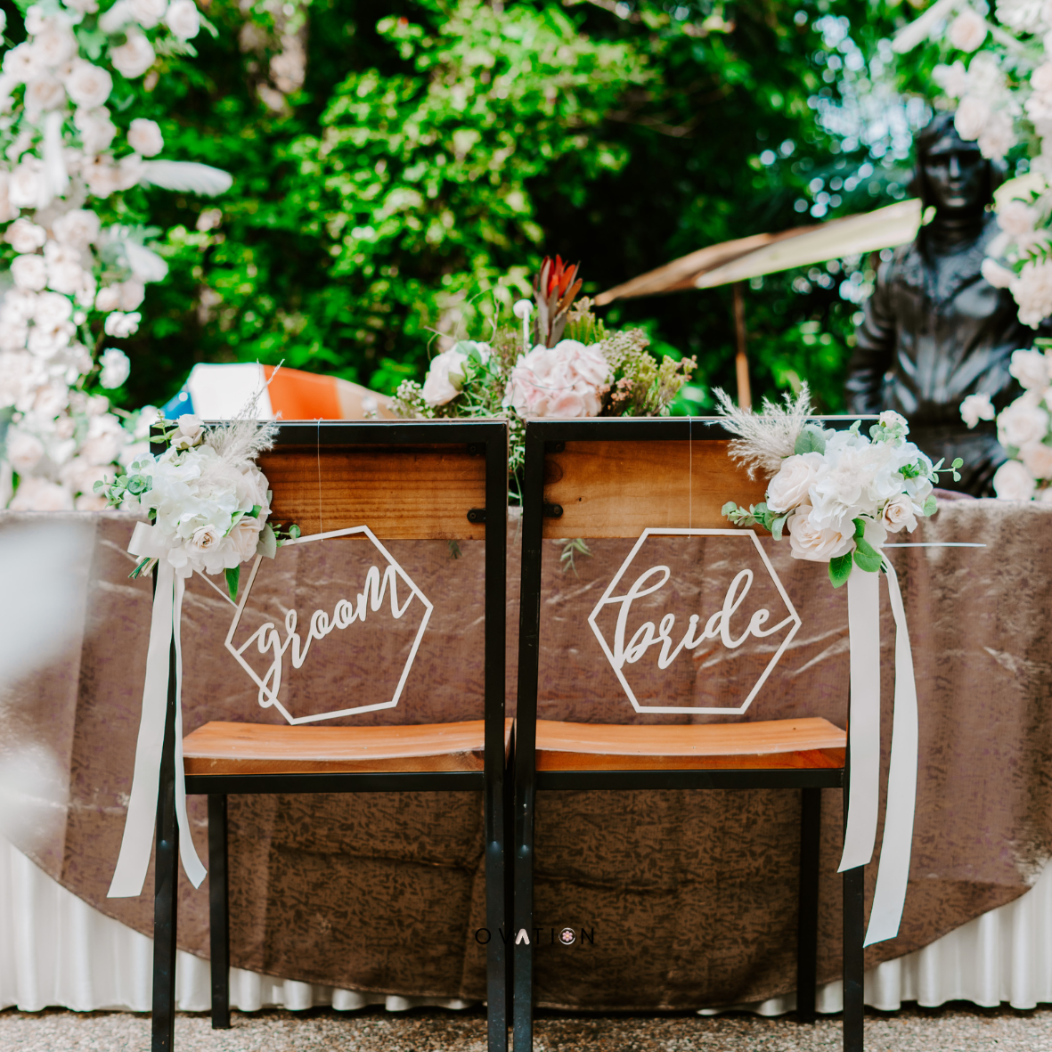 Decorated wedding chairs labeled 'groom' and 'bride' with floral arrangements in an outdoor setting.