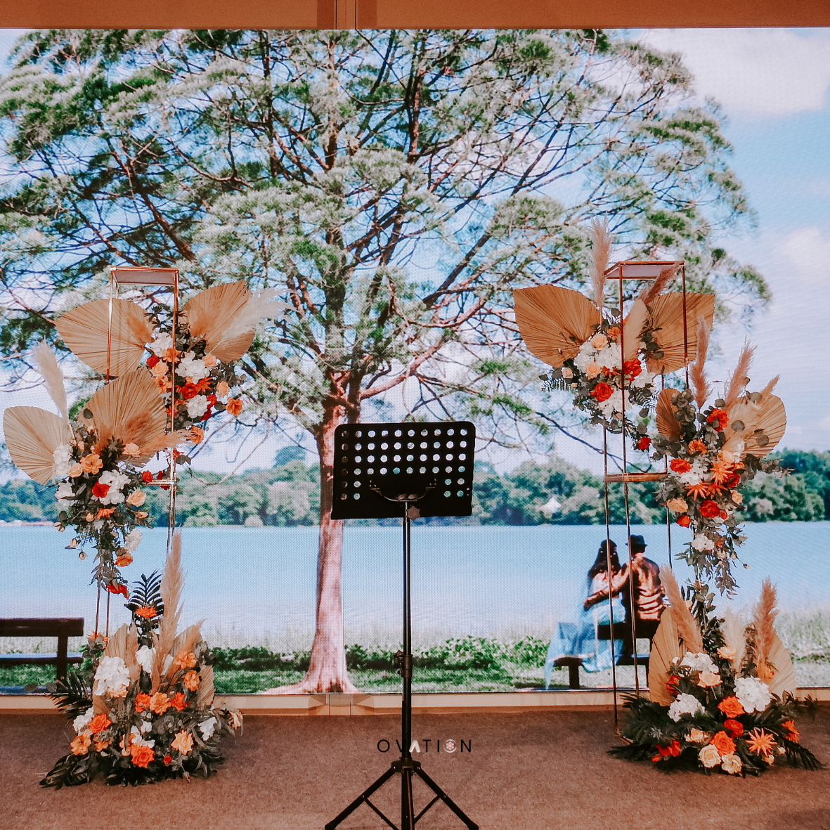 Wedding or event stage with floral arch and backdrop of a large tree by a lake, with two people sitting on a bench in the background.