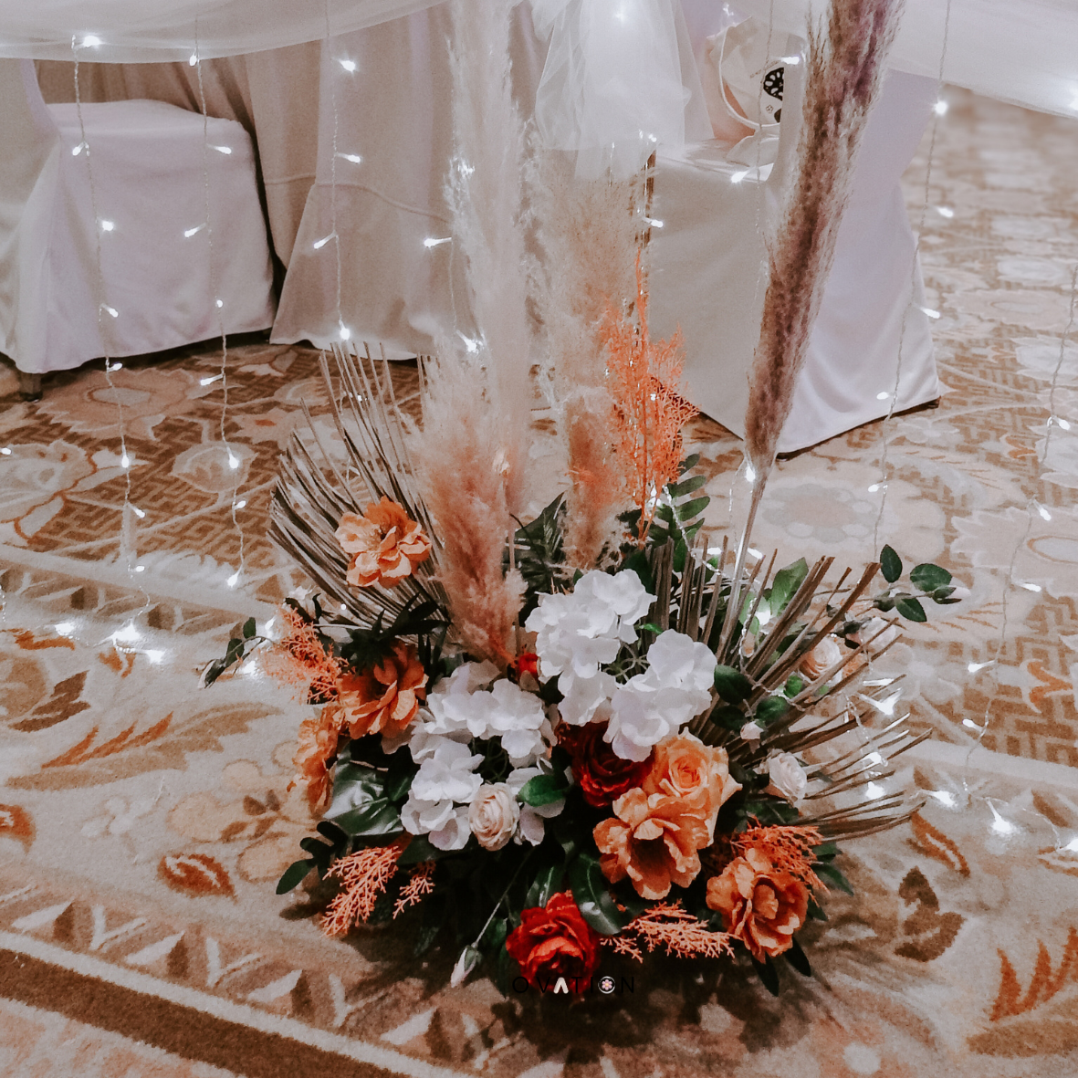 Floral arrangement with orange, white, and red flowers, greenery, and pampas grass, set on a patterned carpet with a backdrop of a table draped in white fabric and string lights.