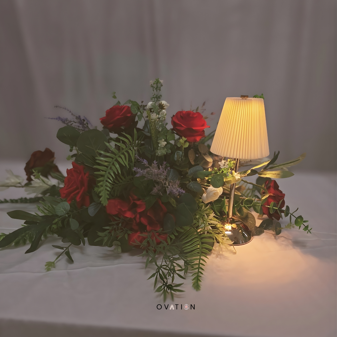 A floral arrangement with red and white roses, greenery, and purple accents, illuminated by a small table lamp with a beige shade, placed on a white surface against a neutral background.