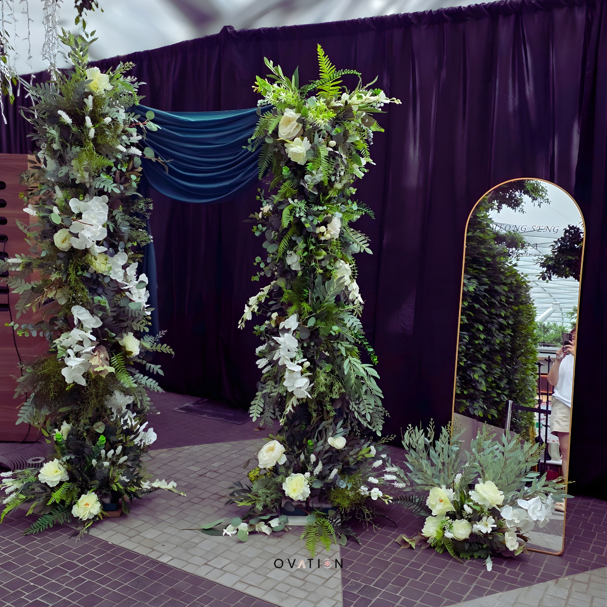 Floral arch with white and green flowers next to a mirror with black curtains in the background.