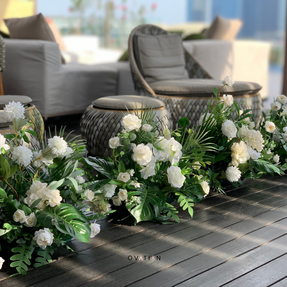 Indoor seating area with wicker chairs and ottomans surrounded by white floral arrangements on a dark wooden floor.
