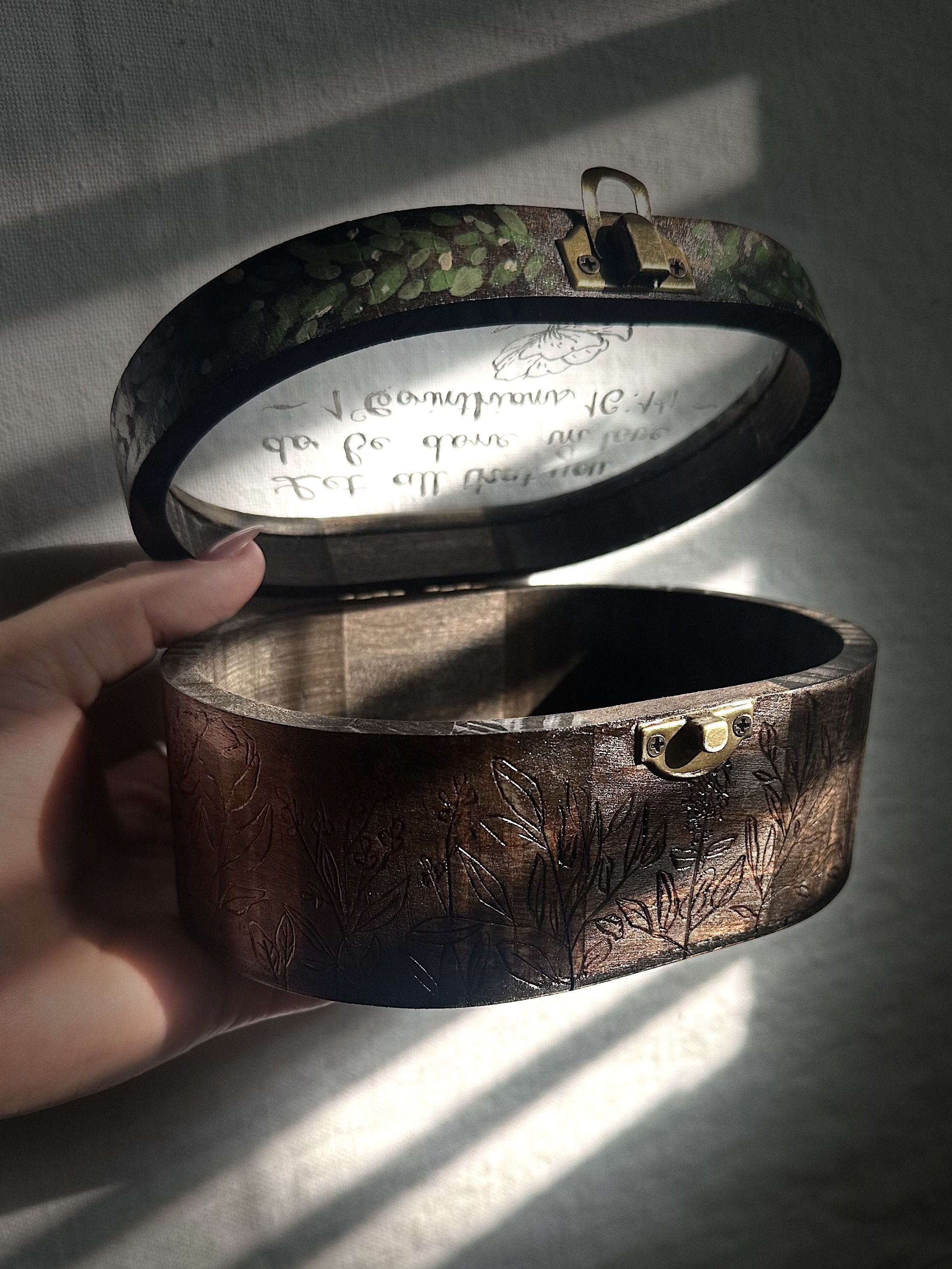 A hand holding a decorative round box with a floral engraved pattern on its outer surface, open to show an inside mirror with a metallic clasp, casting shadows on a light-colored fabric background.