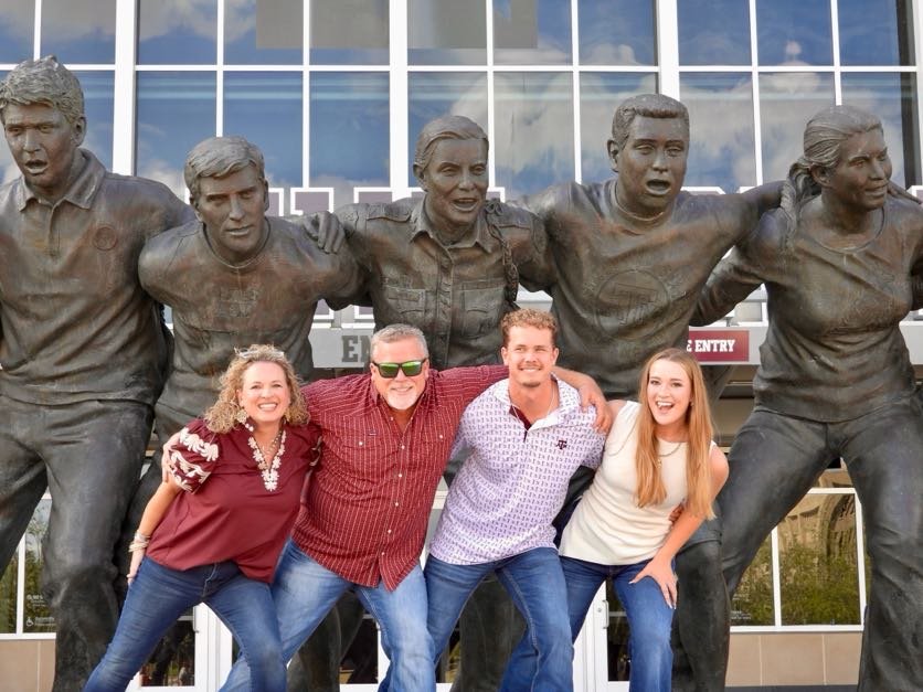Four people smiling and posing in front of a statue of five football players outside a building with large glass windows.