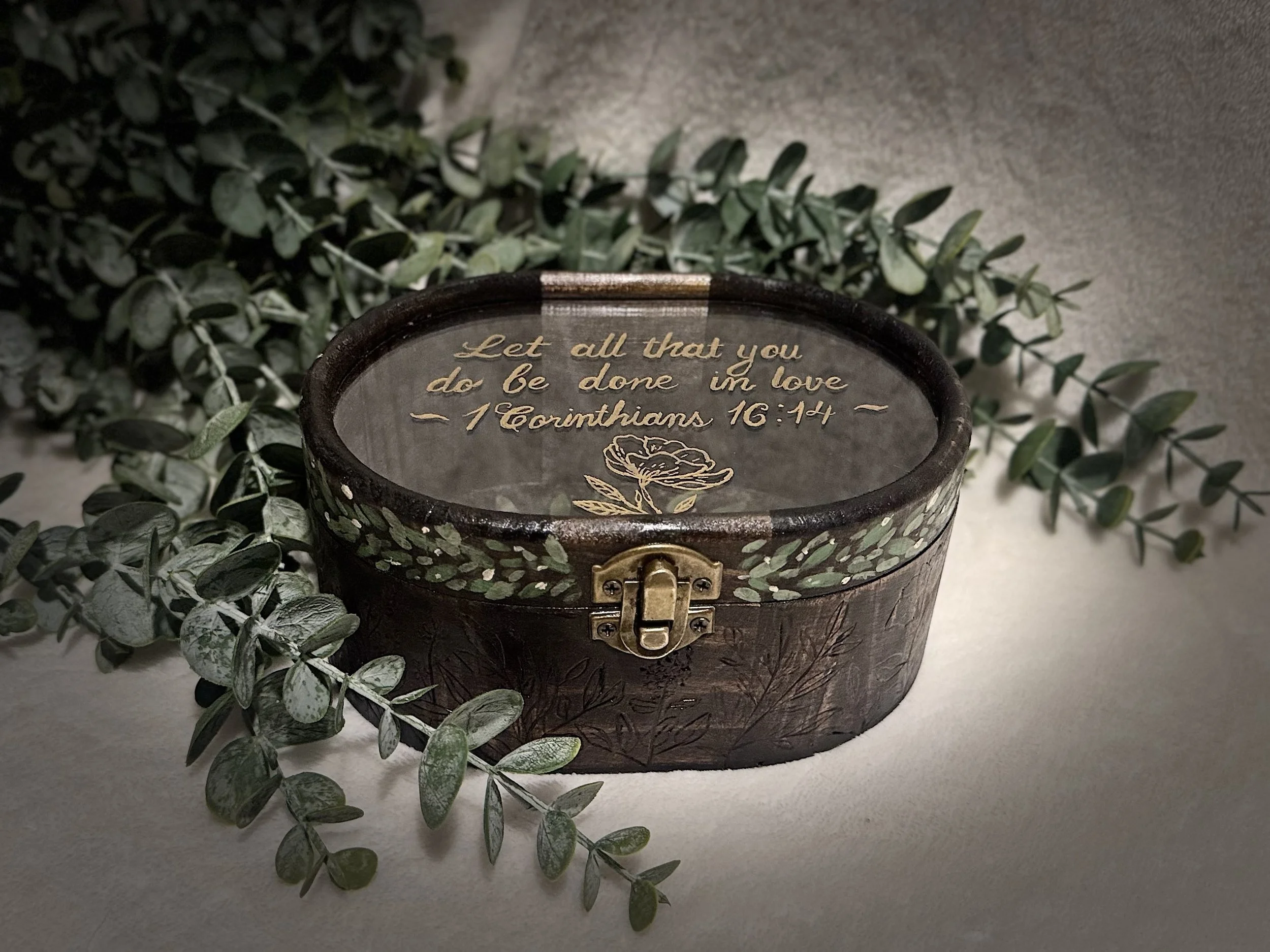 A decorative round box with gold writing on the top that says, "Let all that you do be done in love - 1 Corinthians 16:14". The box has a latch on the front. The background includes green foliage with silver-tinted leaves and a beige surface.