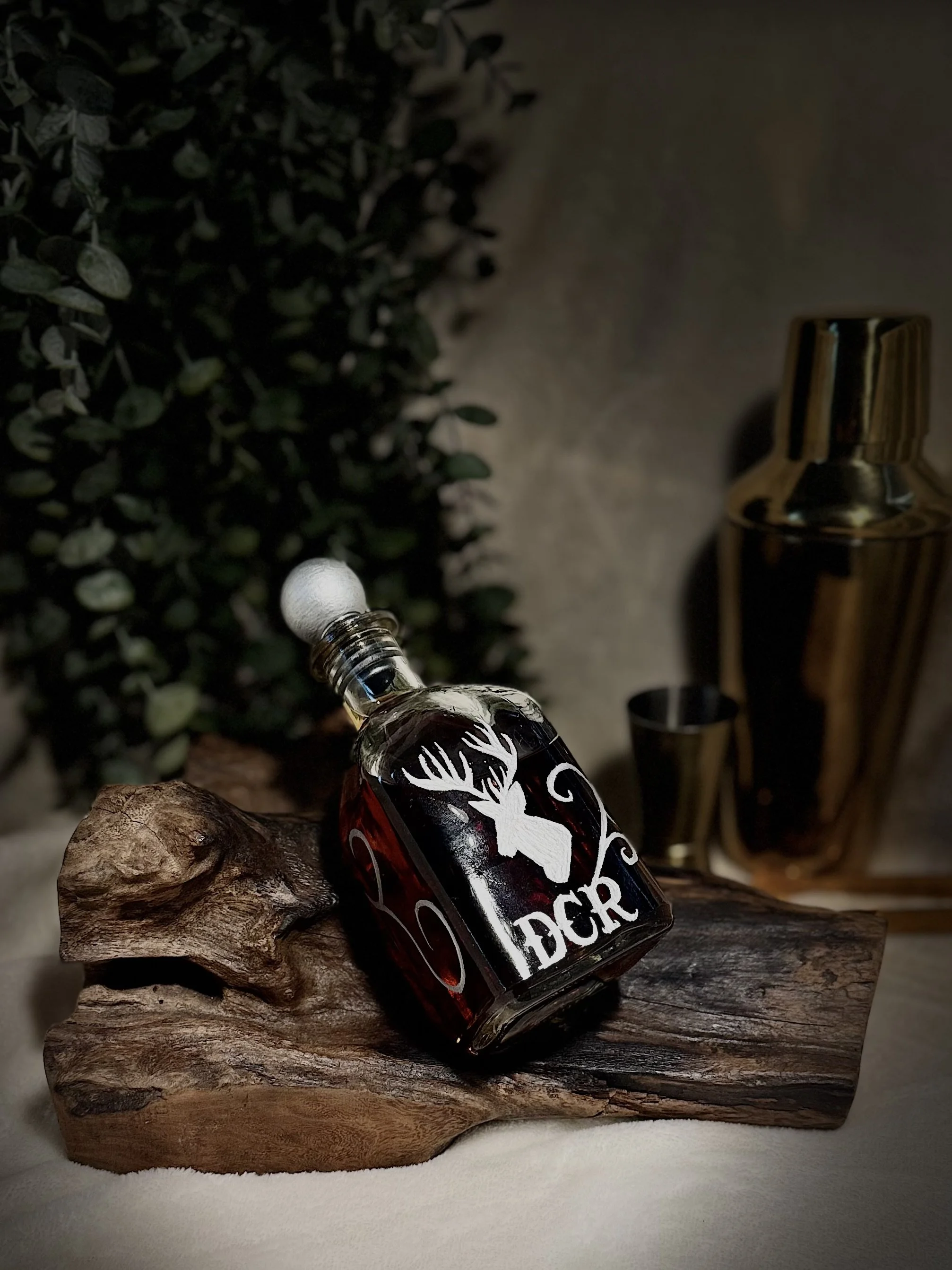 A small bottle labeled 'Hunter' with a white silhouette of a deer, resting on a piece of driftwood on a textured surface. A green shrub, a gold-colored candle holder, and a black cup are in the background.
