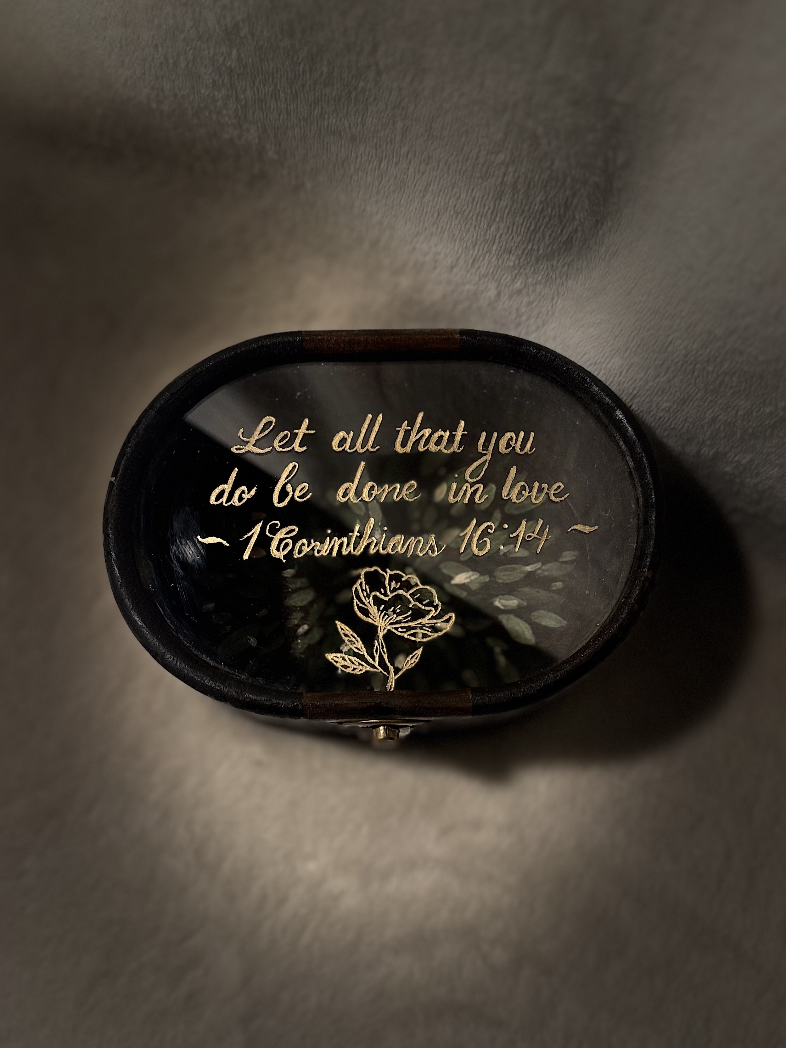 A decorative item with gold foil writing that reads, 'Let all that you do be done in love - 1 Corinthians 16:14,' and features a gold floral design, inside a black frame.