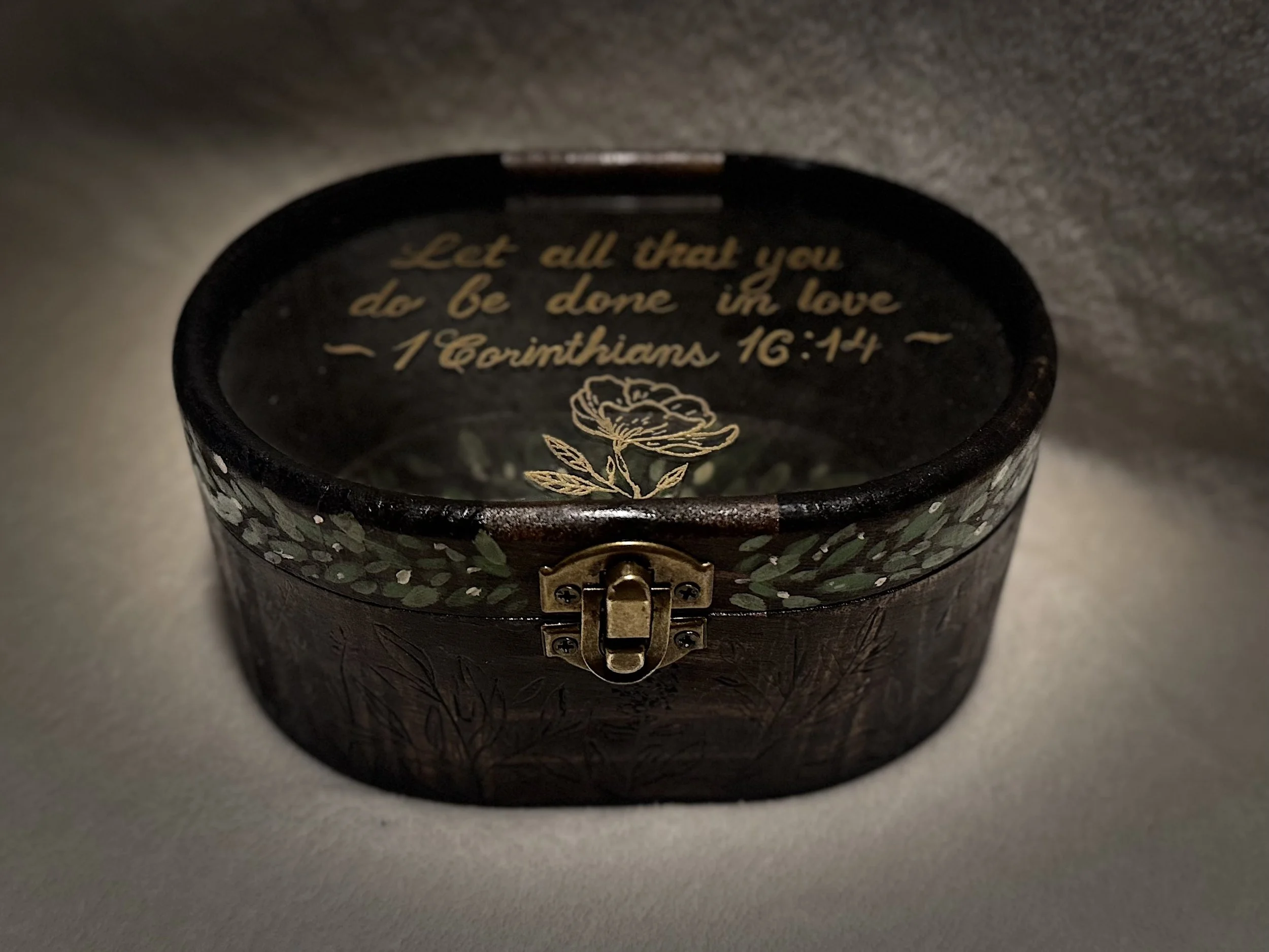 A decorative round box with a hinge and latch, featuring a quote from 1 Corinthians 16:14, and an illustration of a flower on the top, surrounded by a leafy pattern.