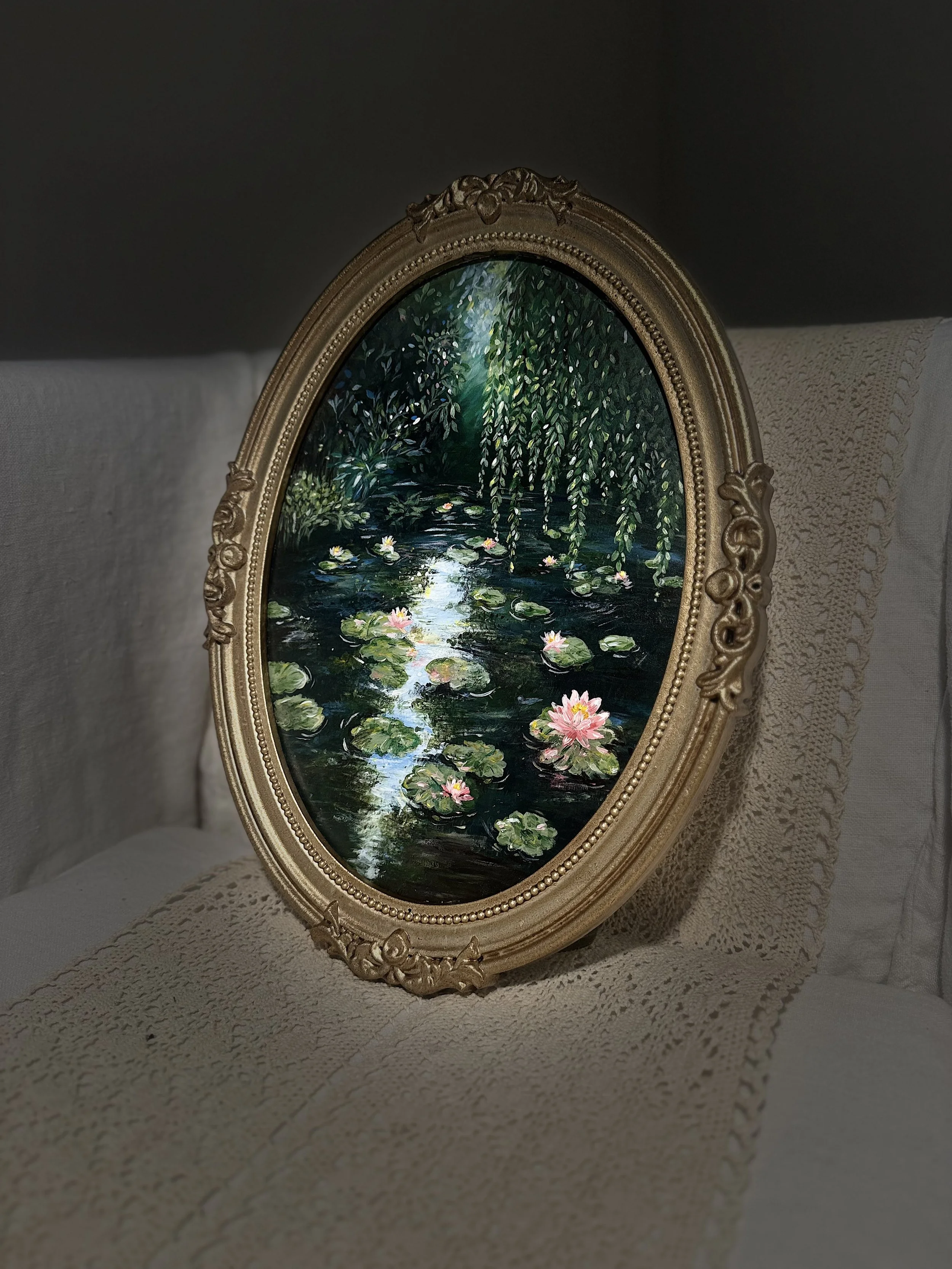 Ornate oval mirror with a painted landscape scene of water lilies and willow trees, placed on a textured off-white surface.
