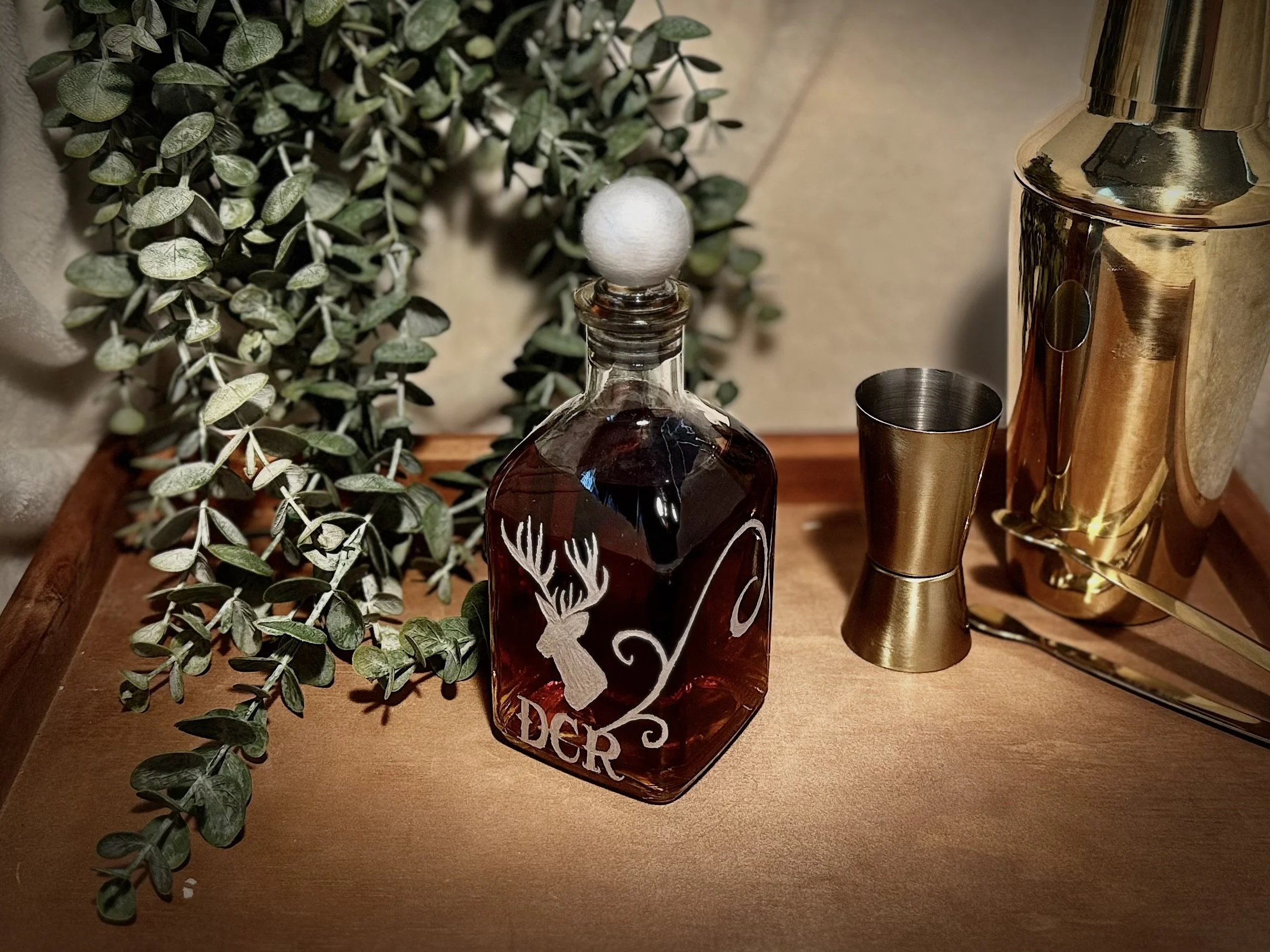 A decorative display featuring a green leafy plant, a brown glass bottle with a white felt ball on top, and a shiny gold-colored cocktail shaker on a wooden surface.