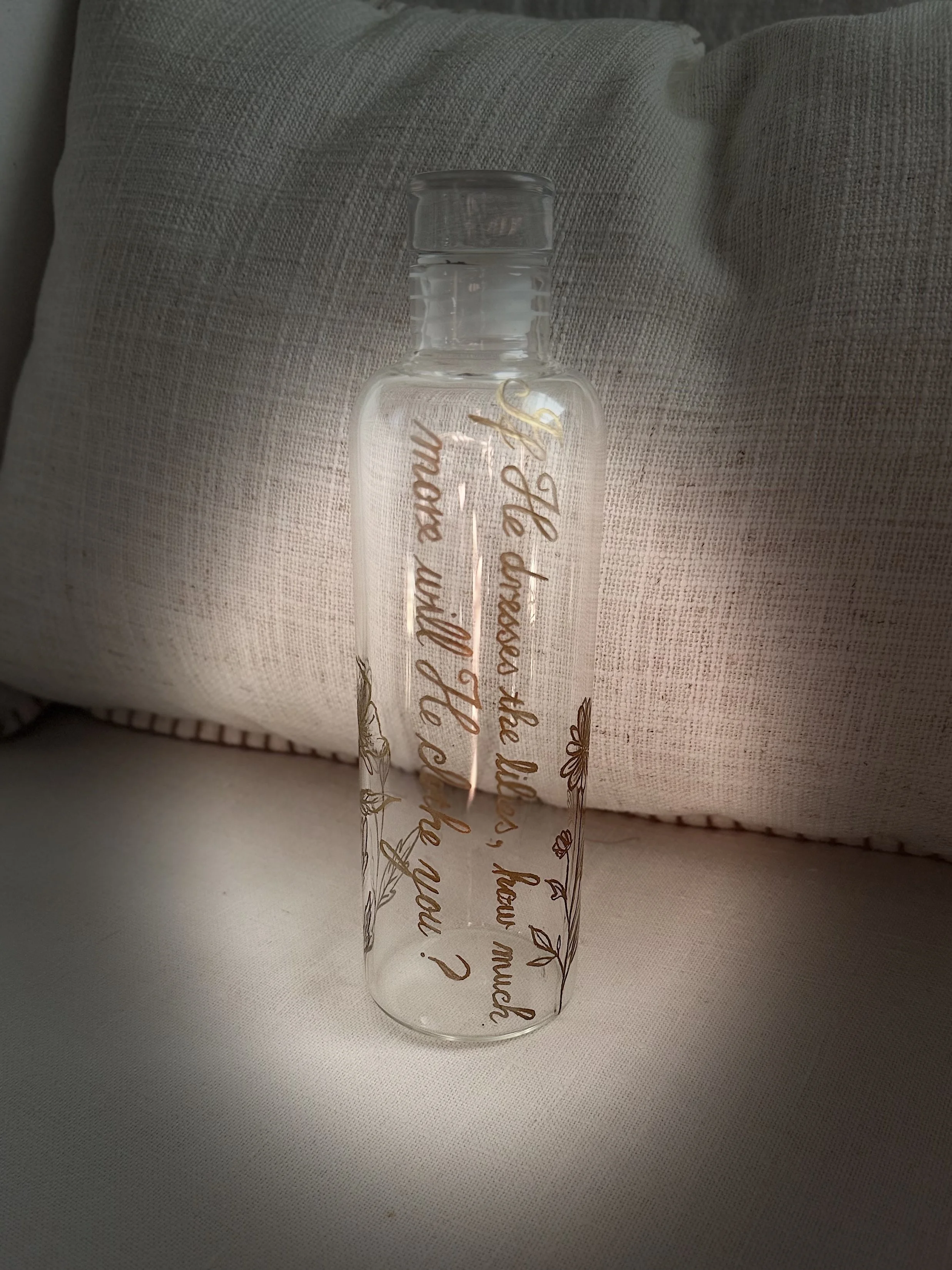 A clear glass bottle with decorative script and floral design, placed on a fabric surface with a pillow in the background.