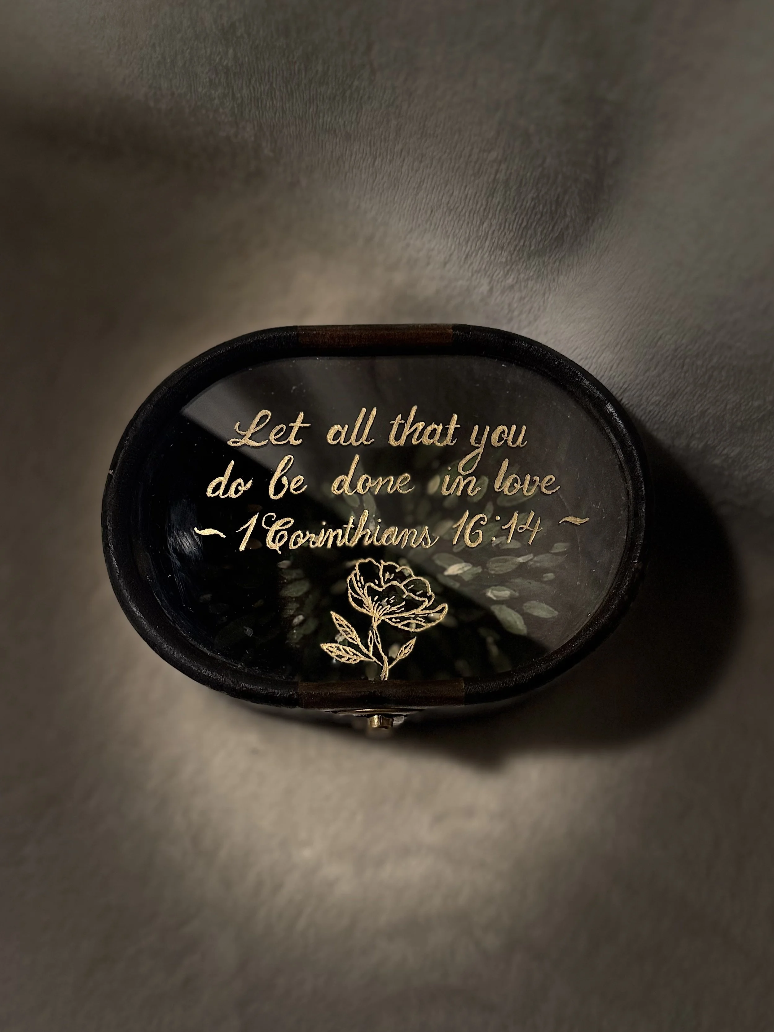 A round black item with a gold inscription of a Bible verse from 1 Corinthians 16:14, and a white floral design, placed on a flat surface.