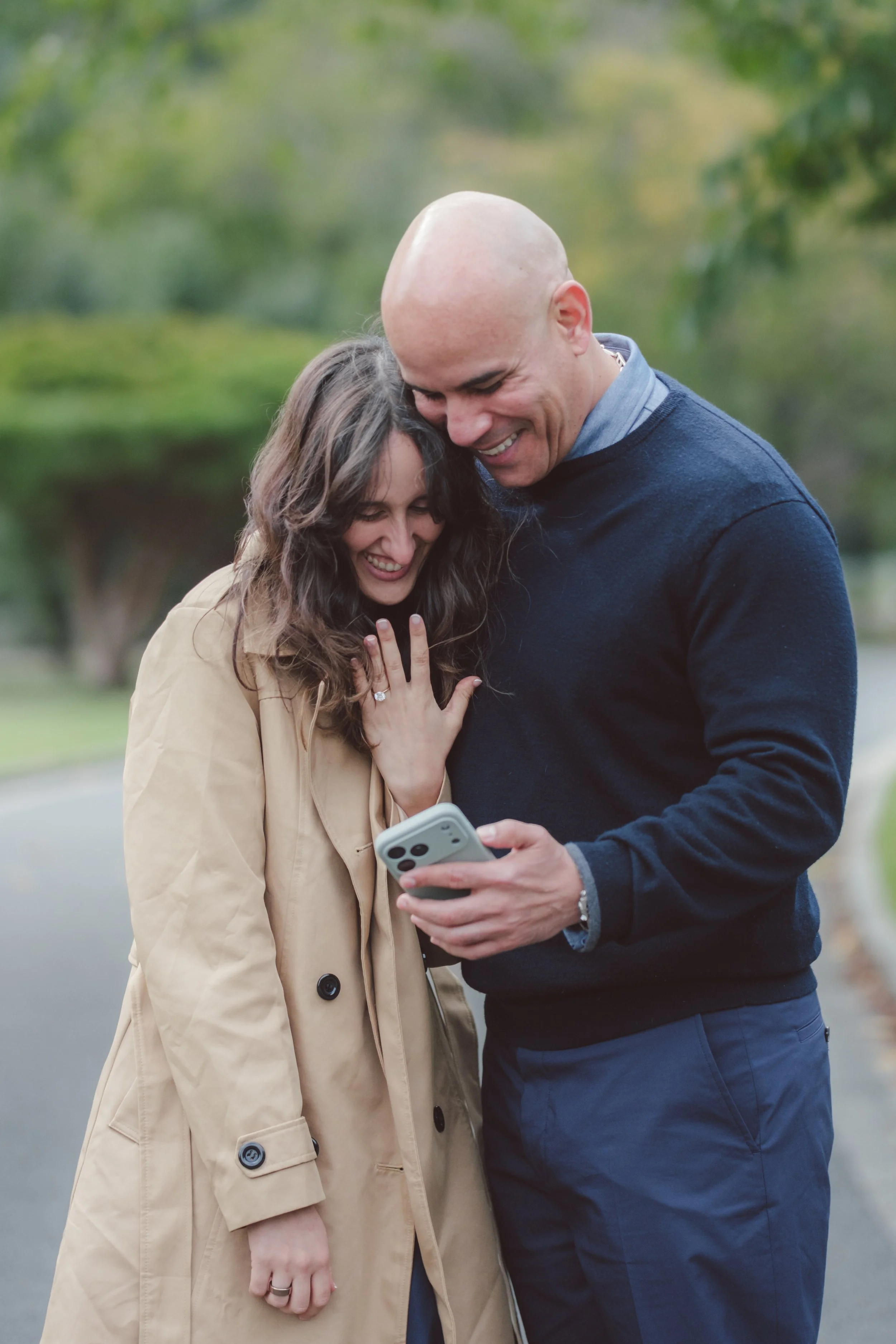 A man and woman are outside walking together, looking at a smartphone, smiling and happy, with green trees and a park in the background.