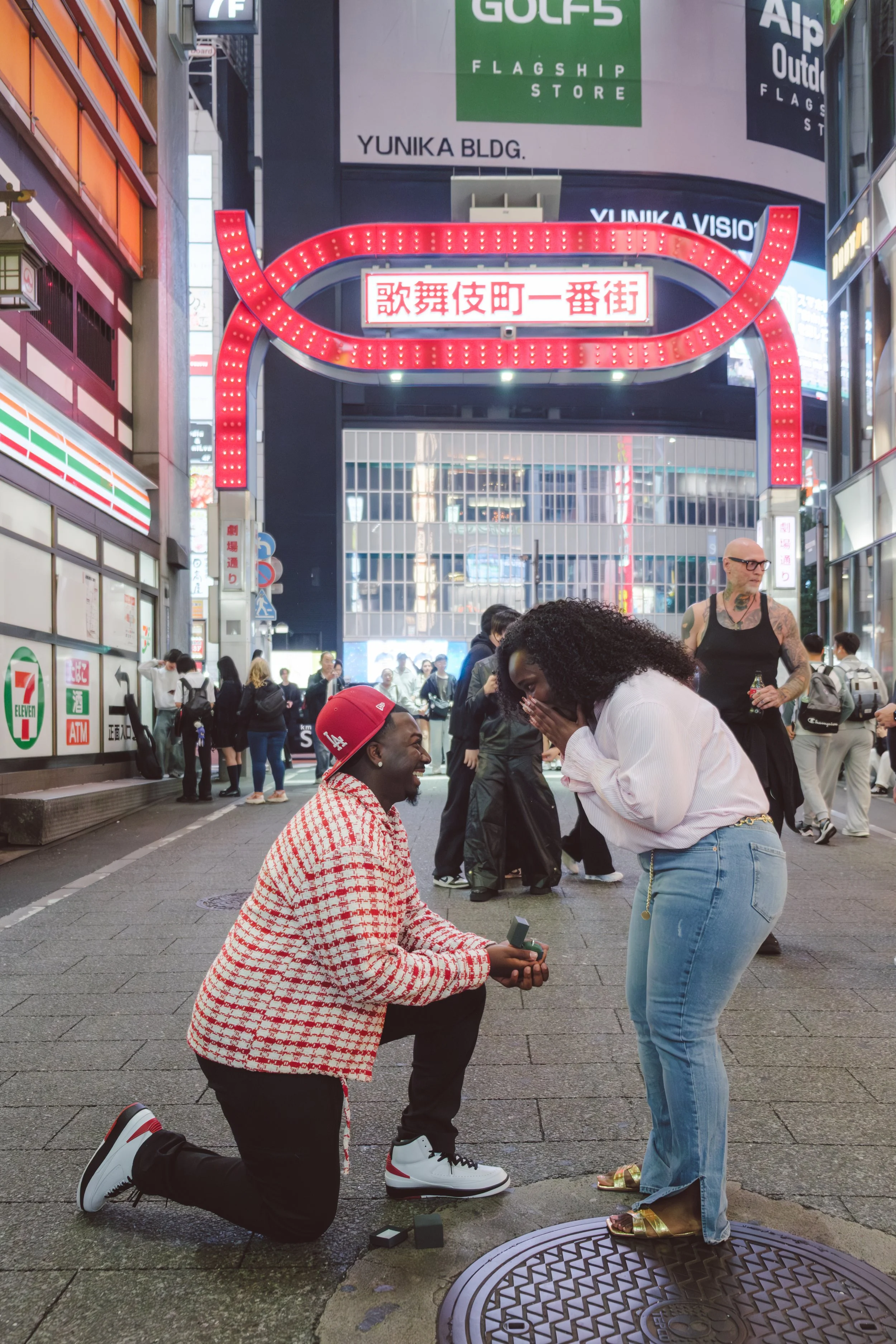 A man is kneeling on one knee proposing marriage to a woman on a busy city street at night. The woman is surprised and covering her mouth with her hands. The man is holding a ring box, and there are people in the background, along with illuminated si