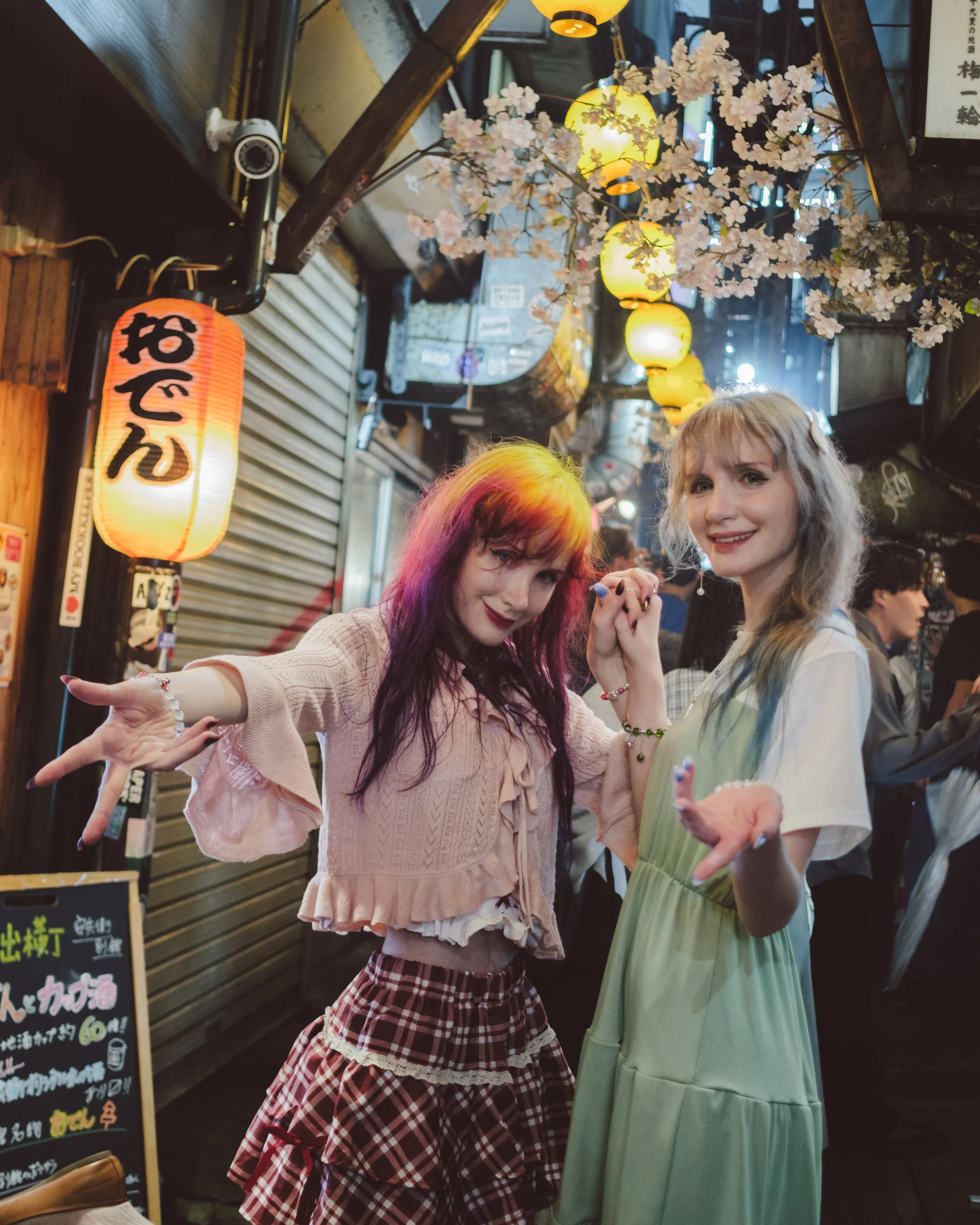 Two young women holding hands and smiling at a night market decorated with cherry blossoms and yellow paper lanterns, with a Japanese sign in the background.