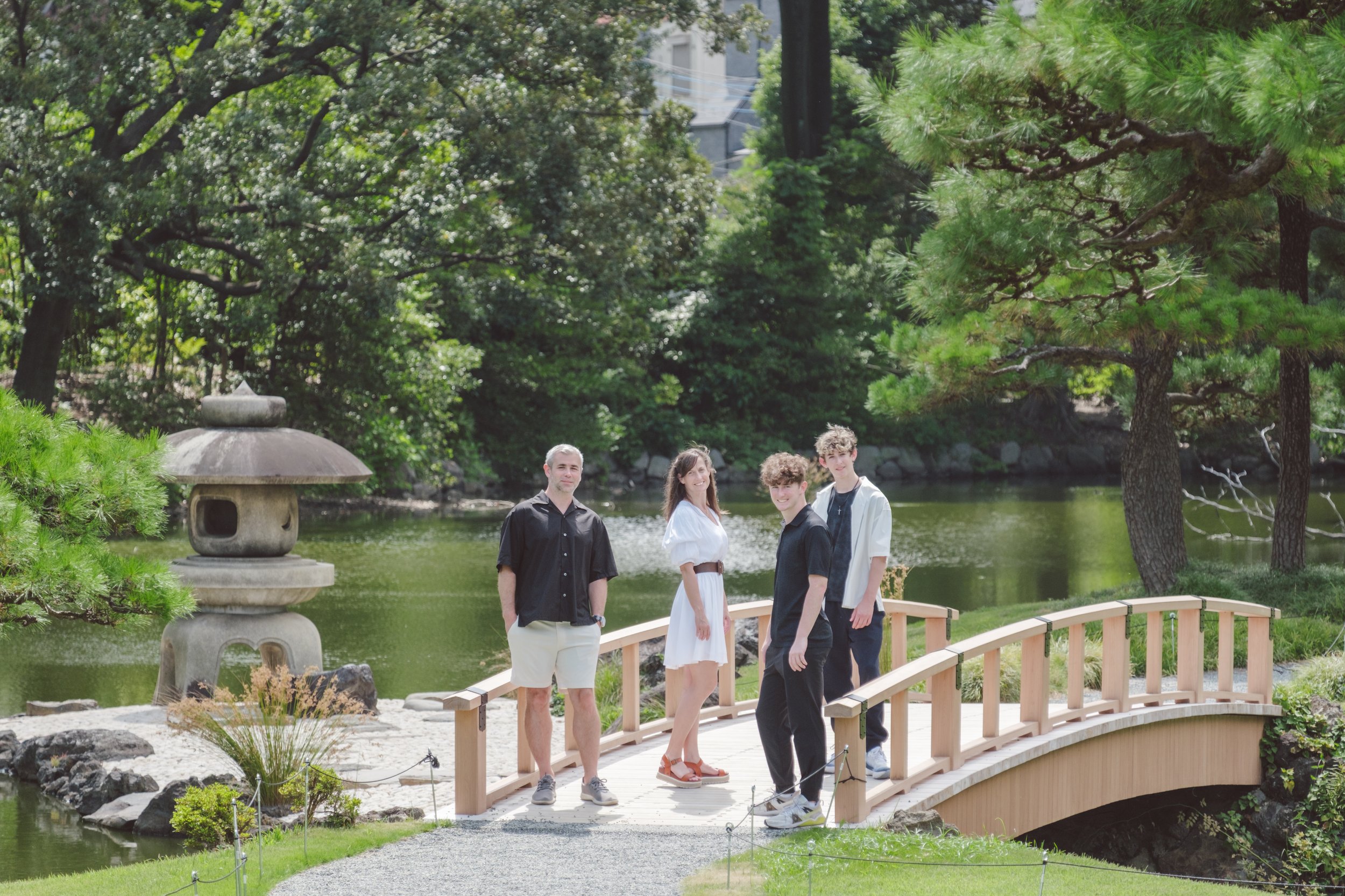 Four young adults stand on a wooden bridge in a Japanese-style garden by a pond, surrounded by lush green trees and rocks, with a stone lantern visible on the left.