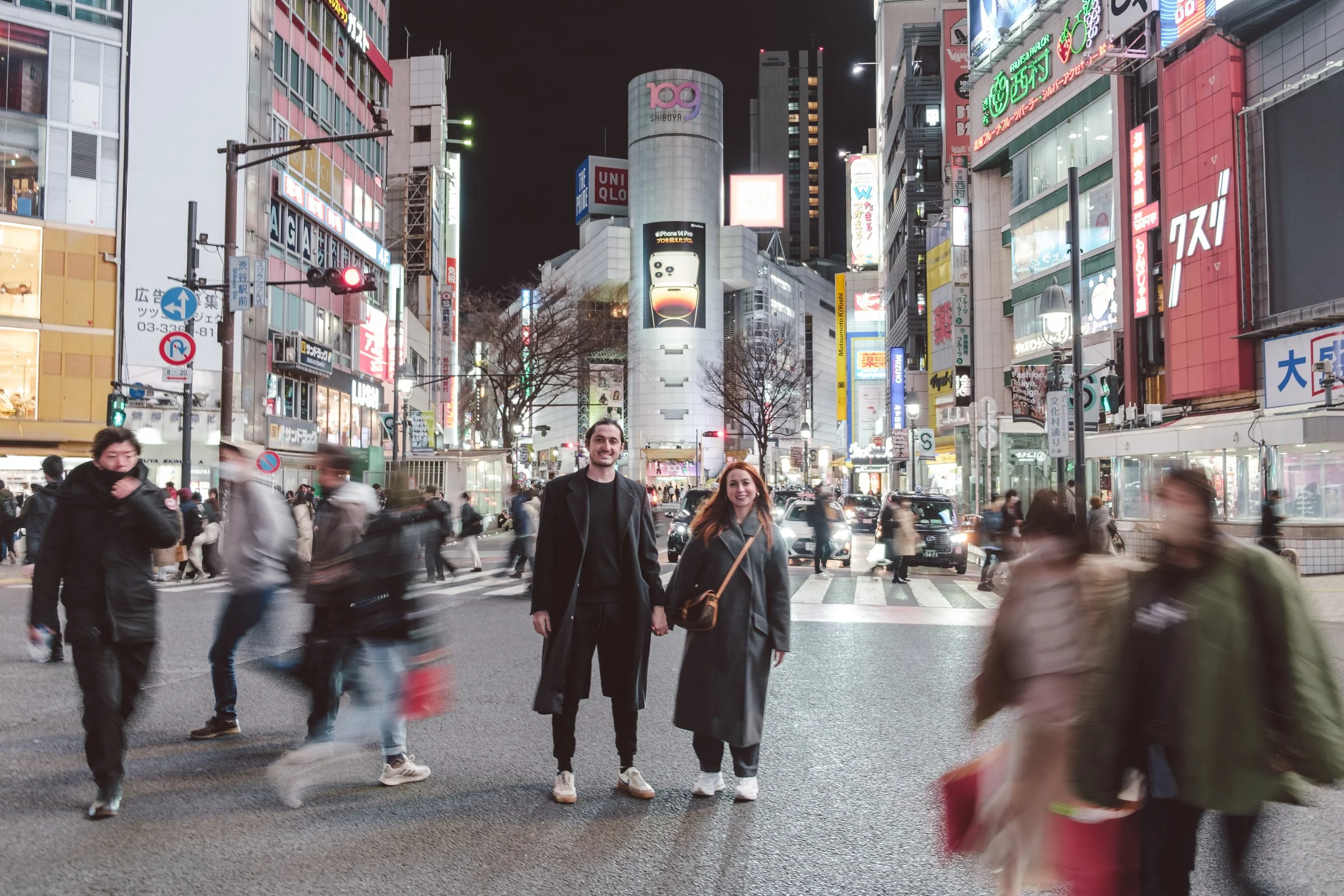 A bustling city street at night with illuminated signs and billboards. Two people are smiling and walking in the crosswalk amidst blurred pedestrians.