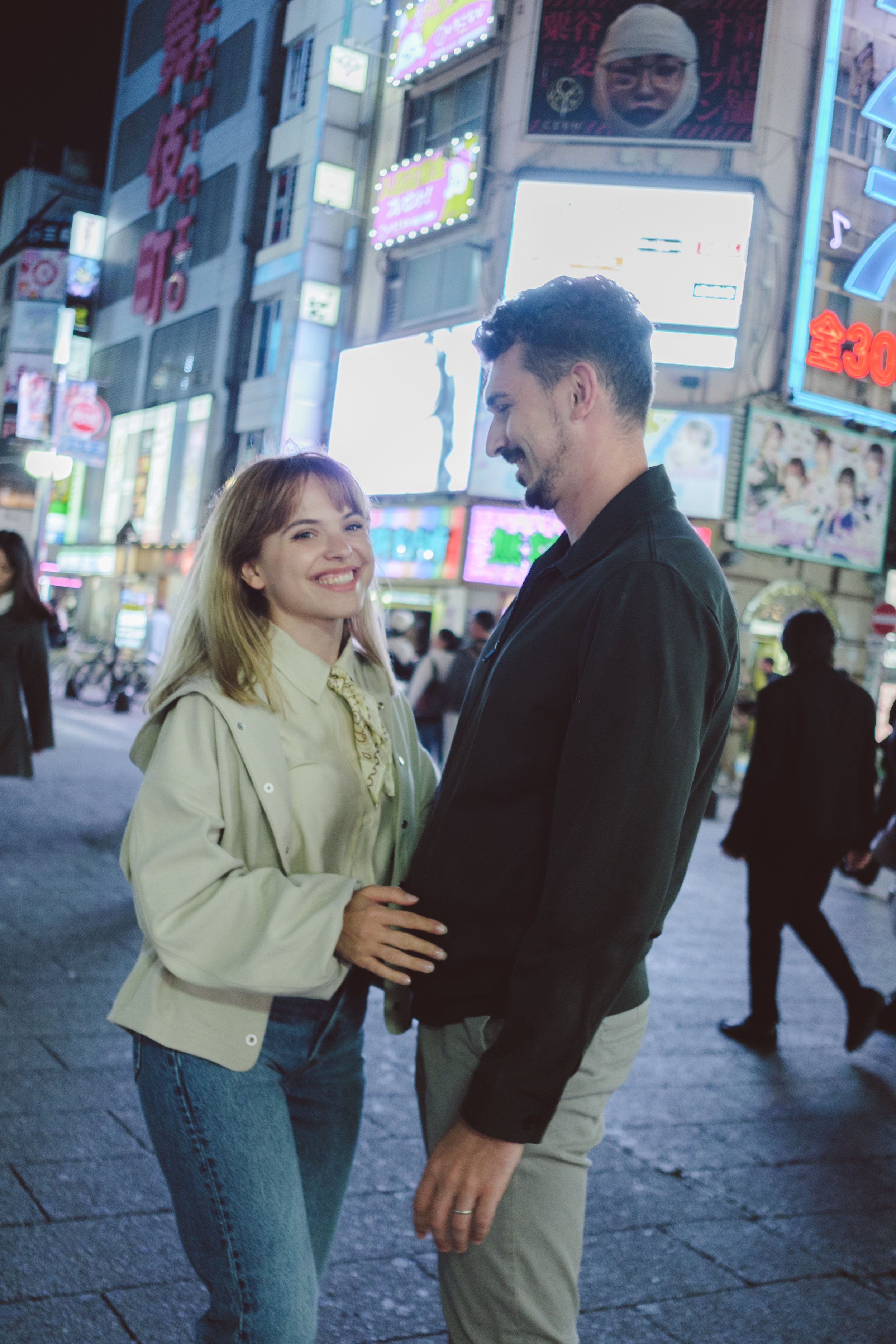 A young woman and a man are holding hands and smiling at each other on a busy city street at night with bright neon signs and billboards in the background.
