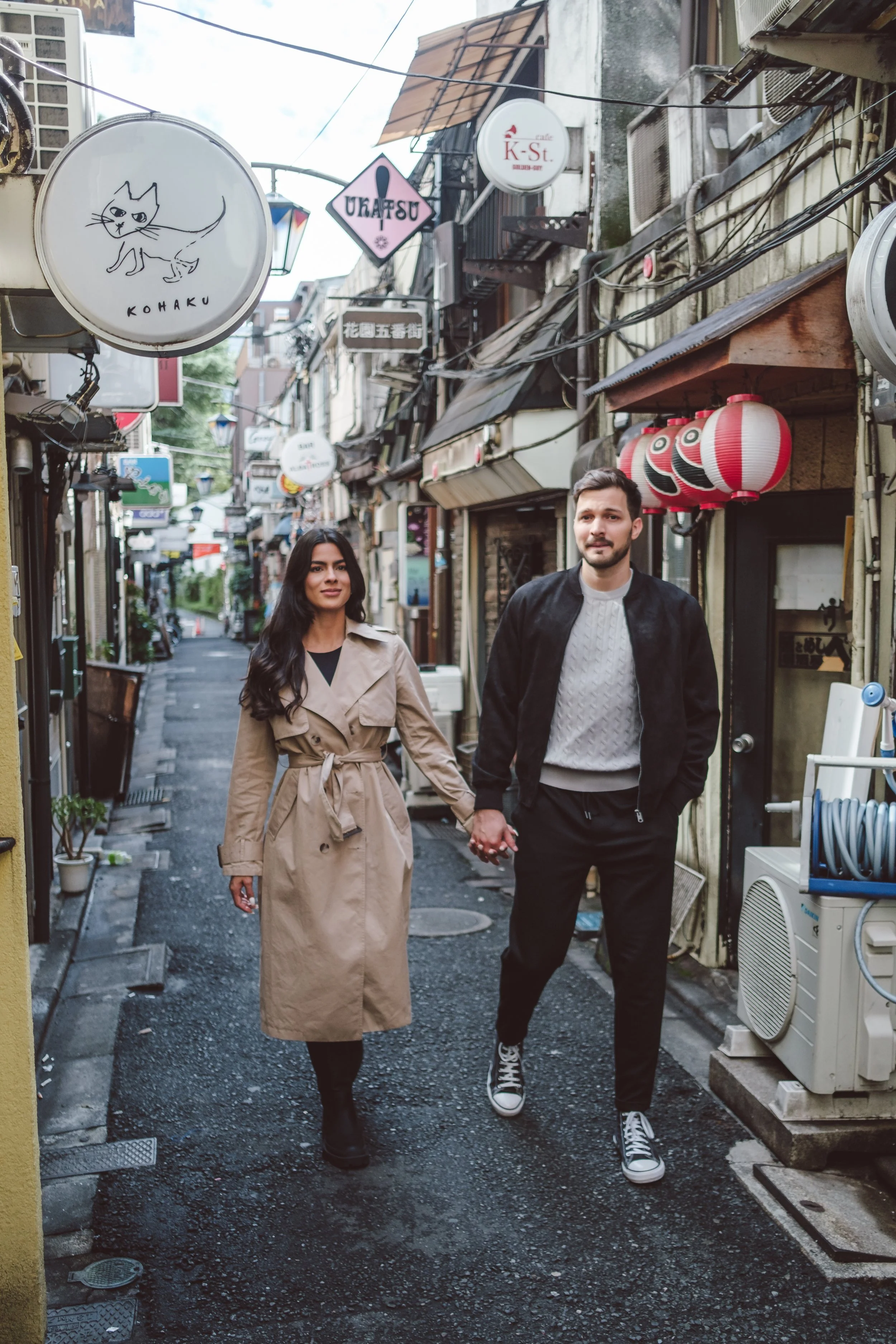 A man and woman walking hand in hand through a narrow alleyway lined with small shops, hanging signs, and traditional lanterns.