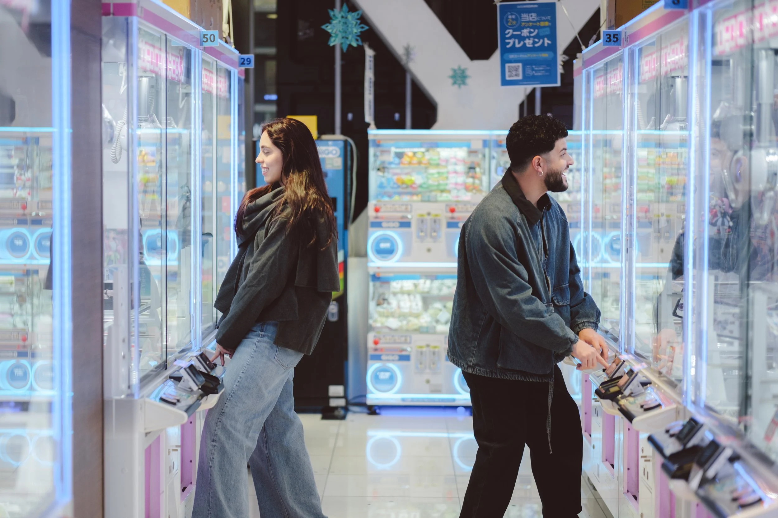 A woman with long brown hair and a man with short dark hair and a beard stand back-to-back in an arcade or gaming center, each facing a different direction and smiling, with rows of claw machines and prize vending machines around them.