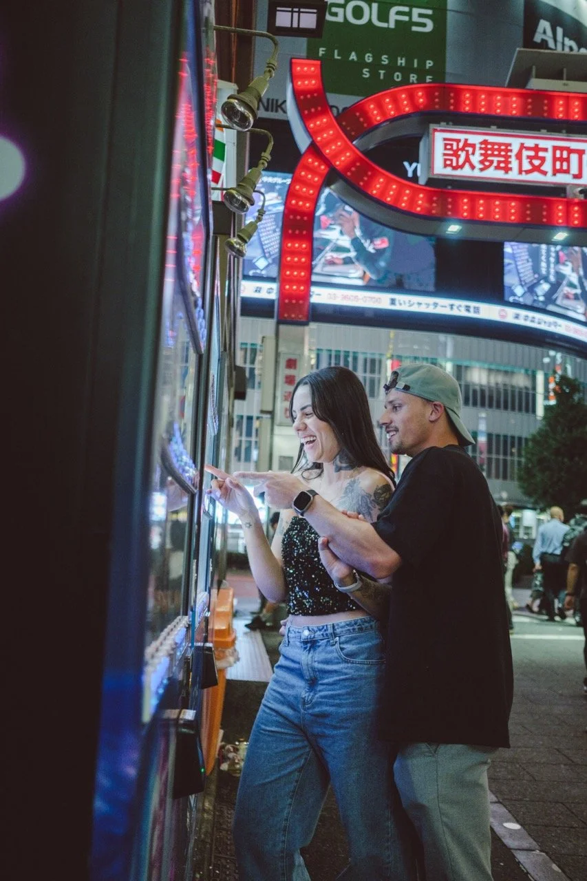 A woman and a man standing next to a row of vending machines on a city street at night, smiling and looking at something on the woman's phone, illuminated by bright lights and neon signs.