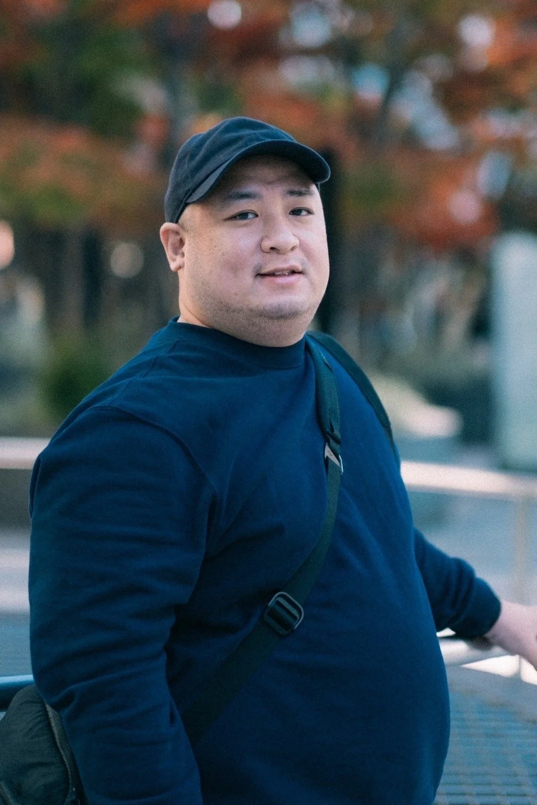A young man with light skin and short dark hair, wearing a black baseball cap and a navy blue sweatshirt, standing outdoors with autumn foliage in the background.