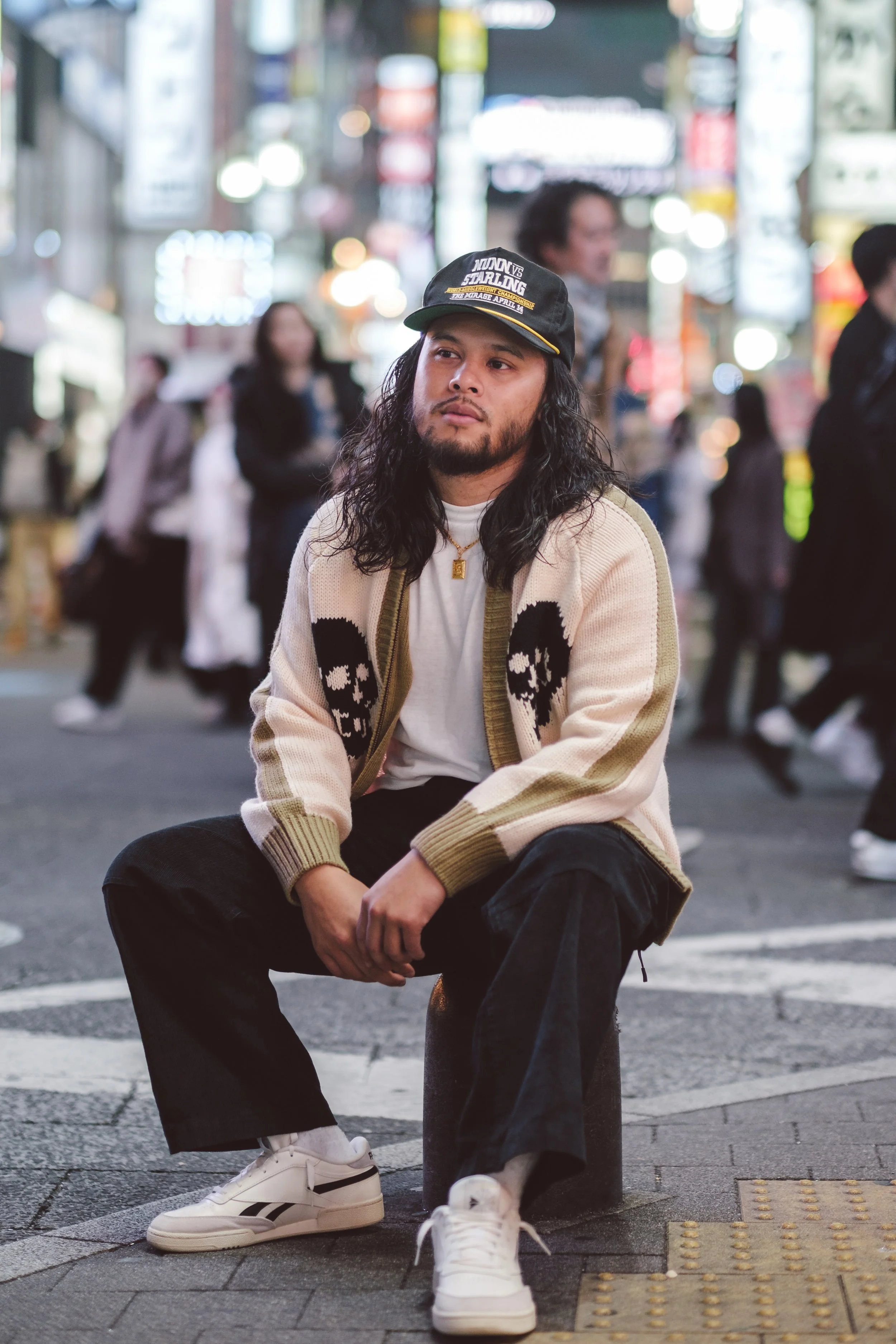 A man with long dark hair and a beard sitting on a stool in a busy city street at night. He is wearing a beige and black sweater with skull designs, black pants, white sneakers, a gold chain necklace, and a black cap with yellow lettering. The backgr