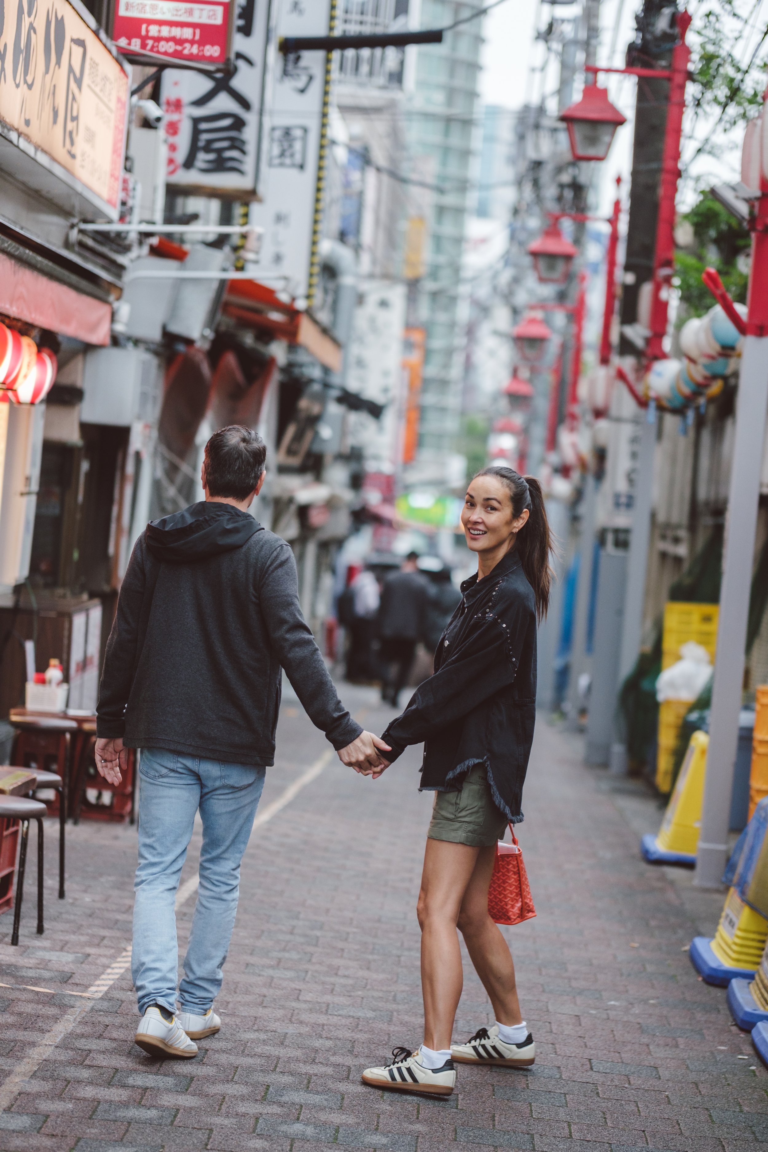 A young woman with dark hair tied in a ponytail, wearing a black jacket, shorts, and Adidas sneakers, holding hands with a man in a black jacket and jeans, walking in an urban street with storefronts, signs, and street lamps.